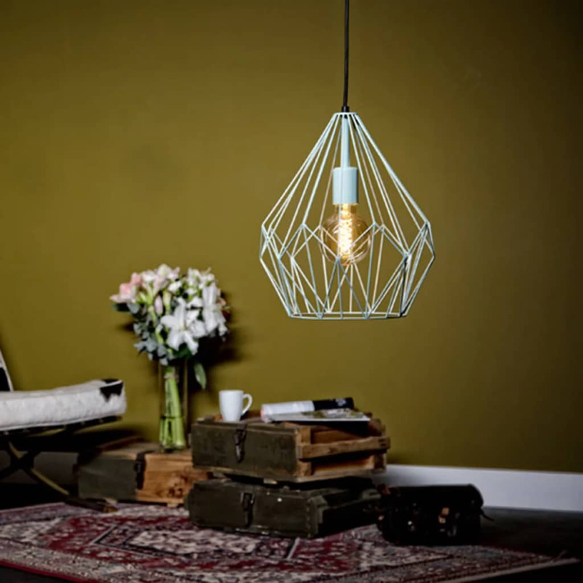 Eine geometrische Pendelleuchte hängt über einer Holzkiste. Eine Vase mit Blumen, eine Tasse und Zeitschriften stehen auf den Kisten. Ein Teppich liegt auf dem Boden.