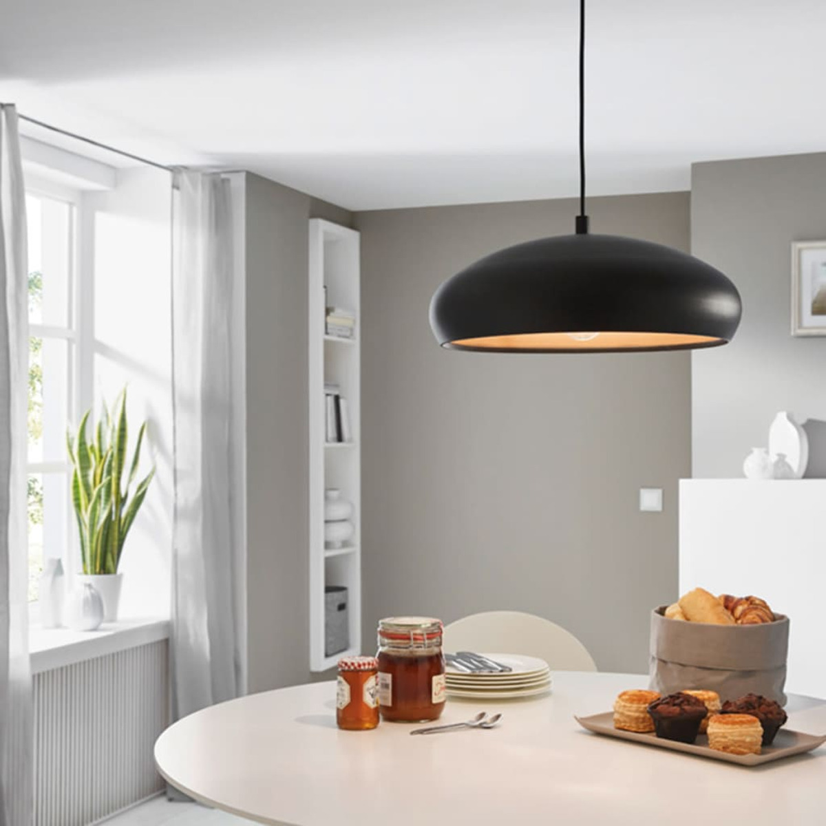 Ein Esszimmer mit einer schwarzen Pendelleuchte über einem weißen Tisch. Marmeladengläser und Gebäck stehen auf dem Tisch. Links ist ein Fenster.