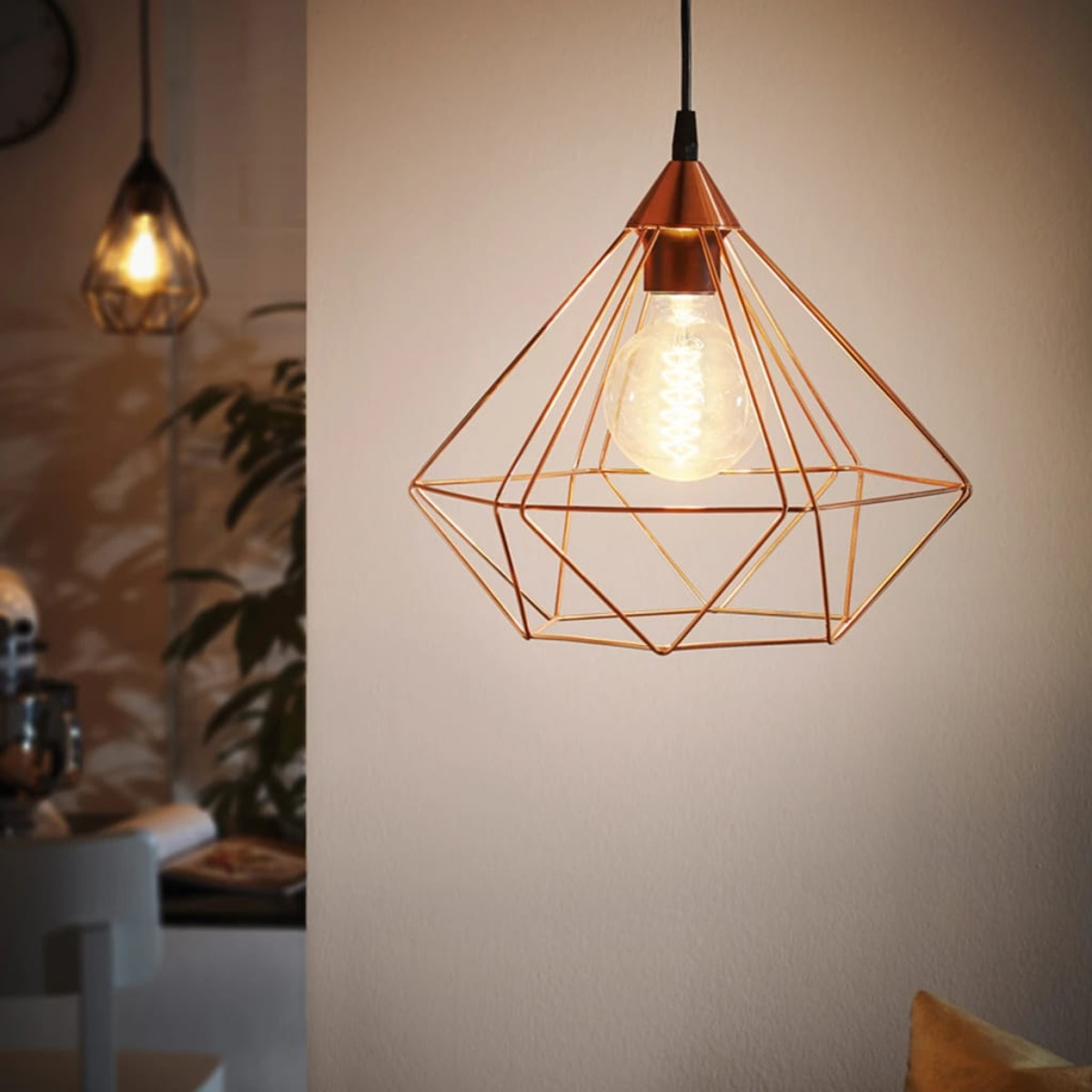 Una lampada a sospensione a forma di diamante in rame con una lampadina accesa pende dal soffitto. Un'altra lampada simile è sullo sfondo sfocato.