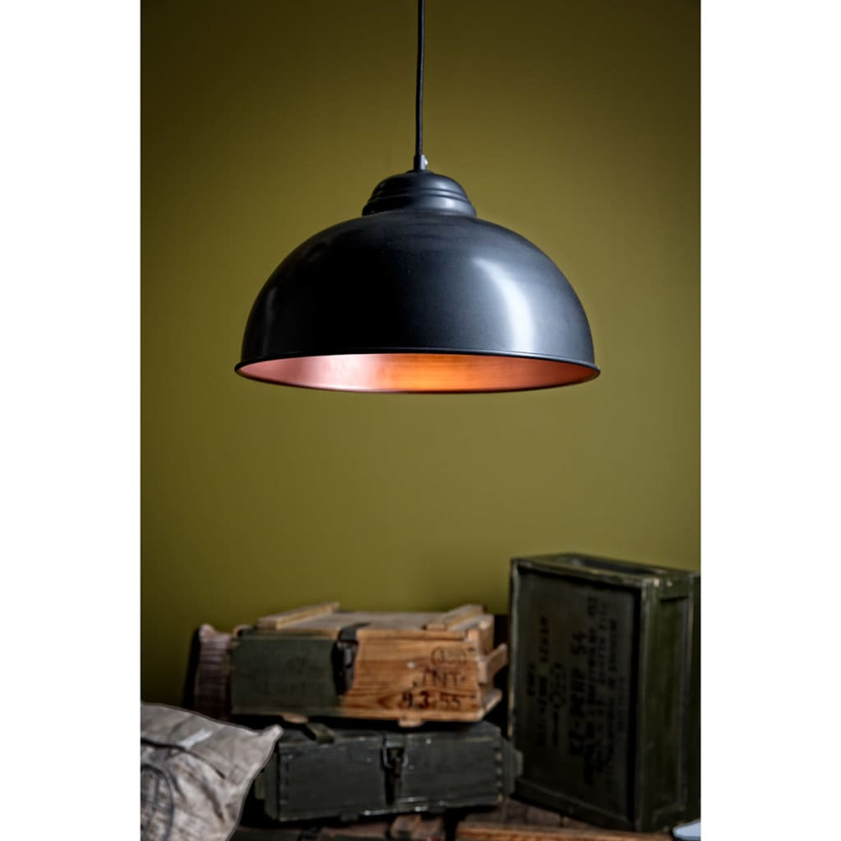 Eine schwarze Hängelampe mit Kupferinnenraum hängt über Holzkisten und einer grünen Wand. Die Lampe ist der zentrale Fokus.