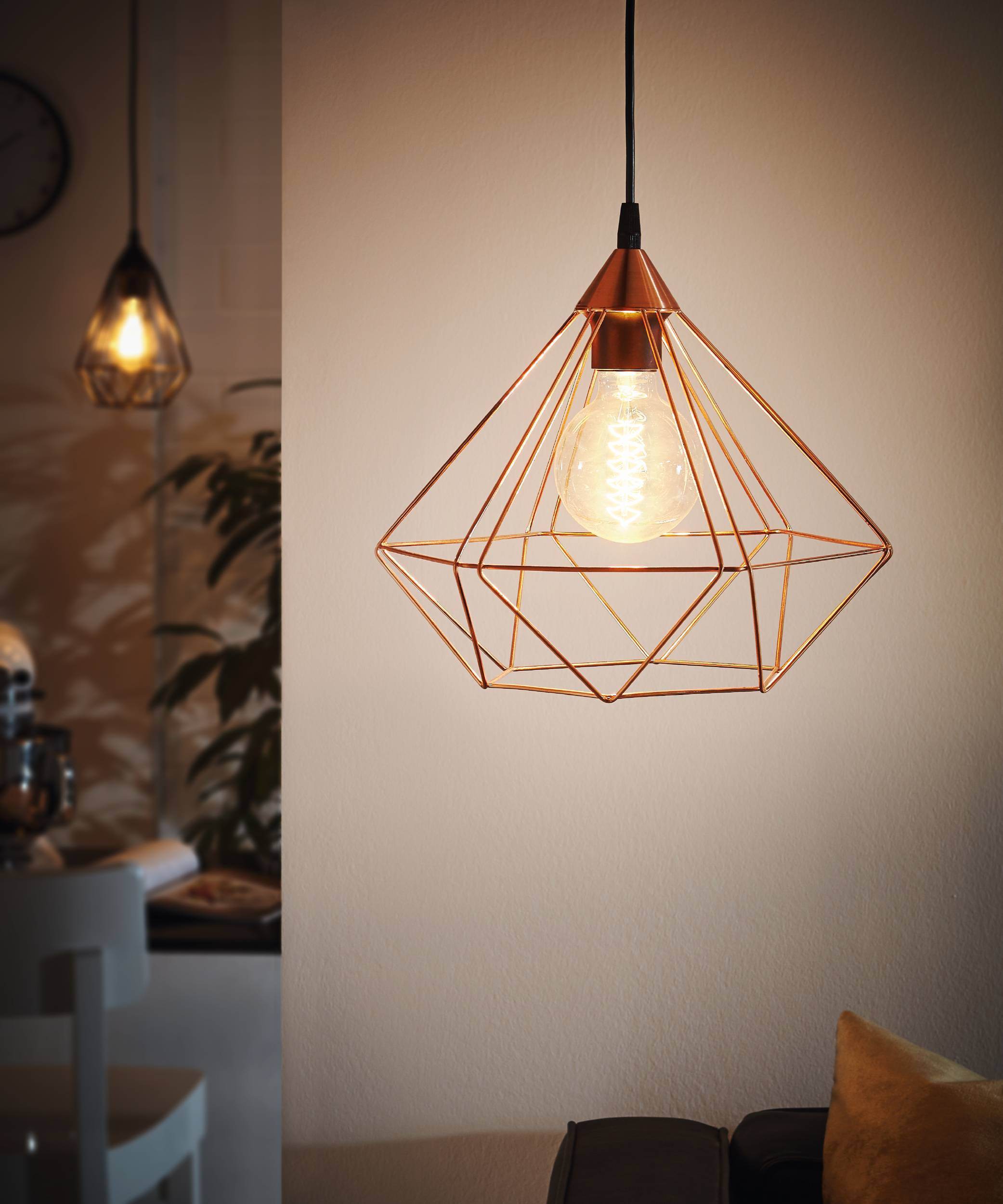 Una lampada a sospensione in rame con gabbia a diamante, che illumina una stanza con una calda luce. Un'altra luce è visibile sullo sfondo.