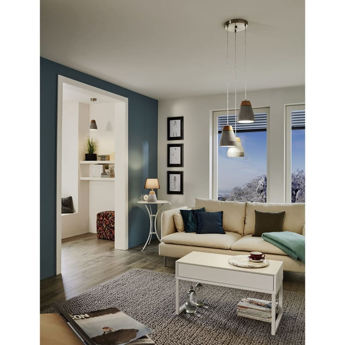 Wohnzimmer mit beigem Sofa, weißem Tisch und drei Hängelampen. Durch die Fenster ist eine schneebedeckte Landschaft zu sehen.