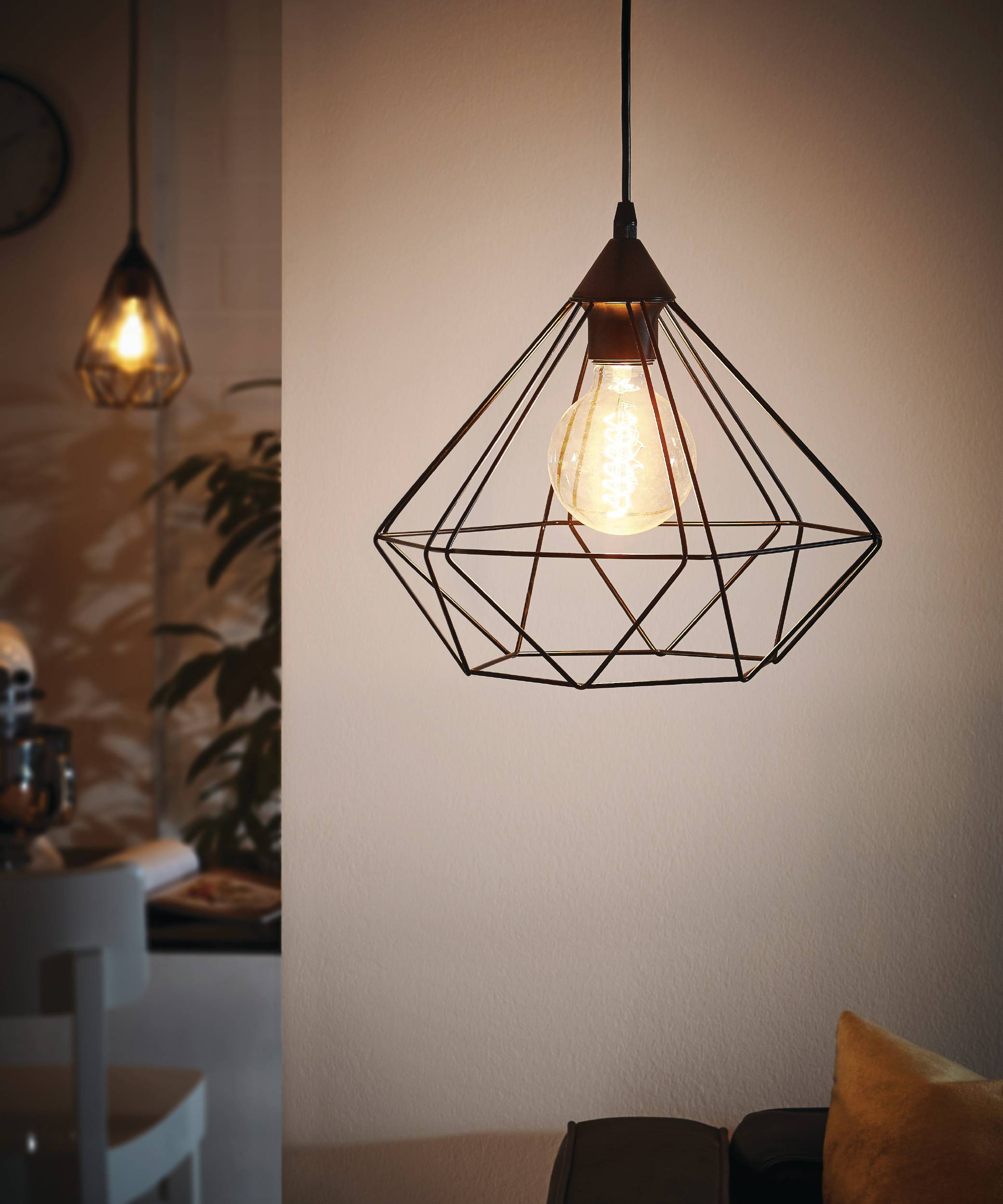 Eine geometrische Pendelleuchte mit einer beleuchteten Glühbirne hängt. Eine weitere Lampe befindet sich im Hintergrund, in der Nähe einer Pflanze.