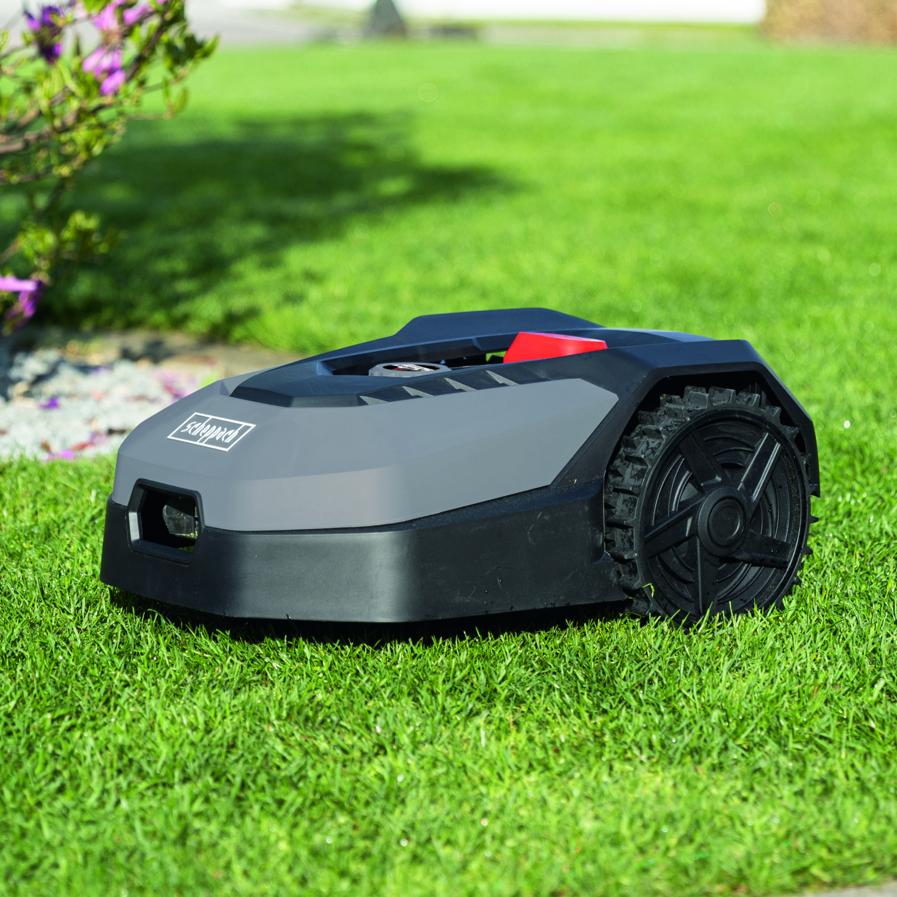 Kosiarka robotyczna na zielonej trawie. Ma szary korpus z czarnymi kołami i czerwonym przyciskiem.