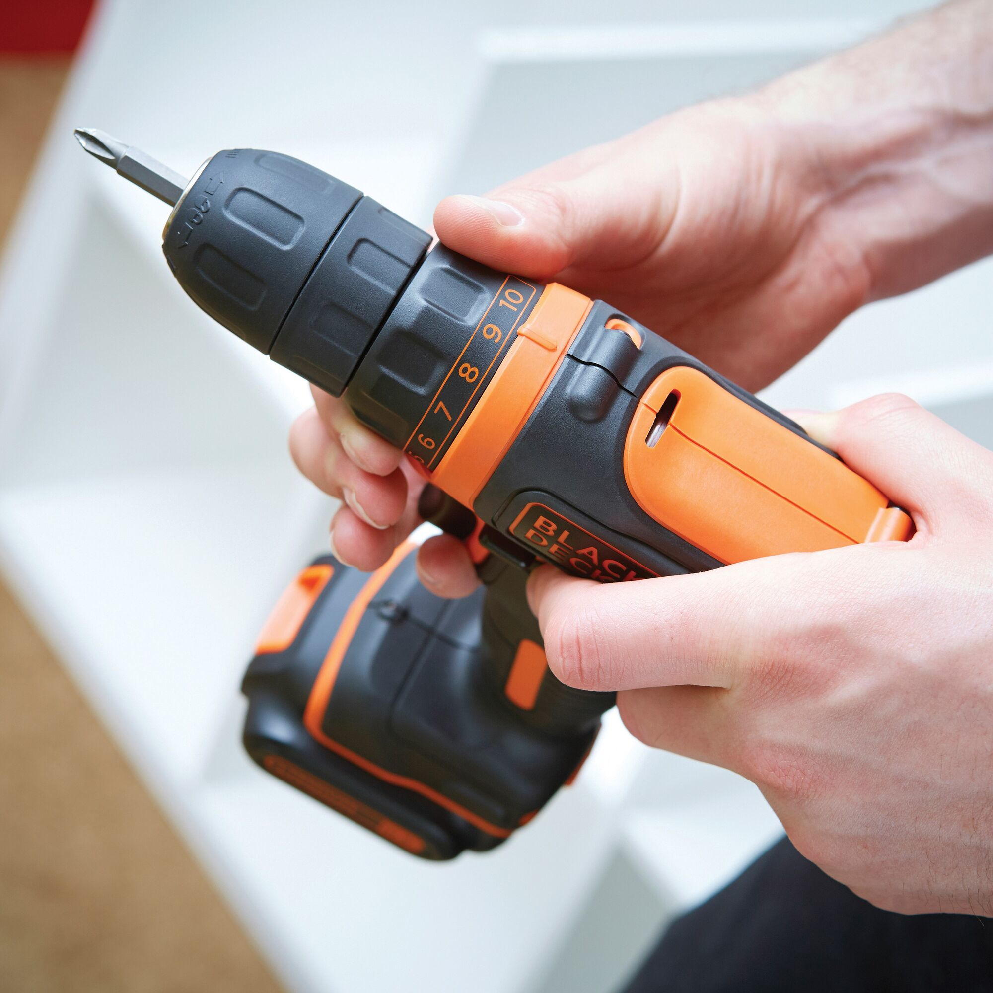 Mani che tengono un trapano avvitatore nero e arancione Black+Decker, con una punta argentata. Scaffali bianchi sullo sfondo.