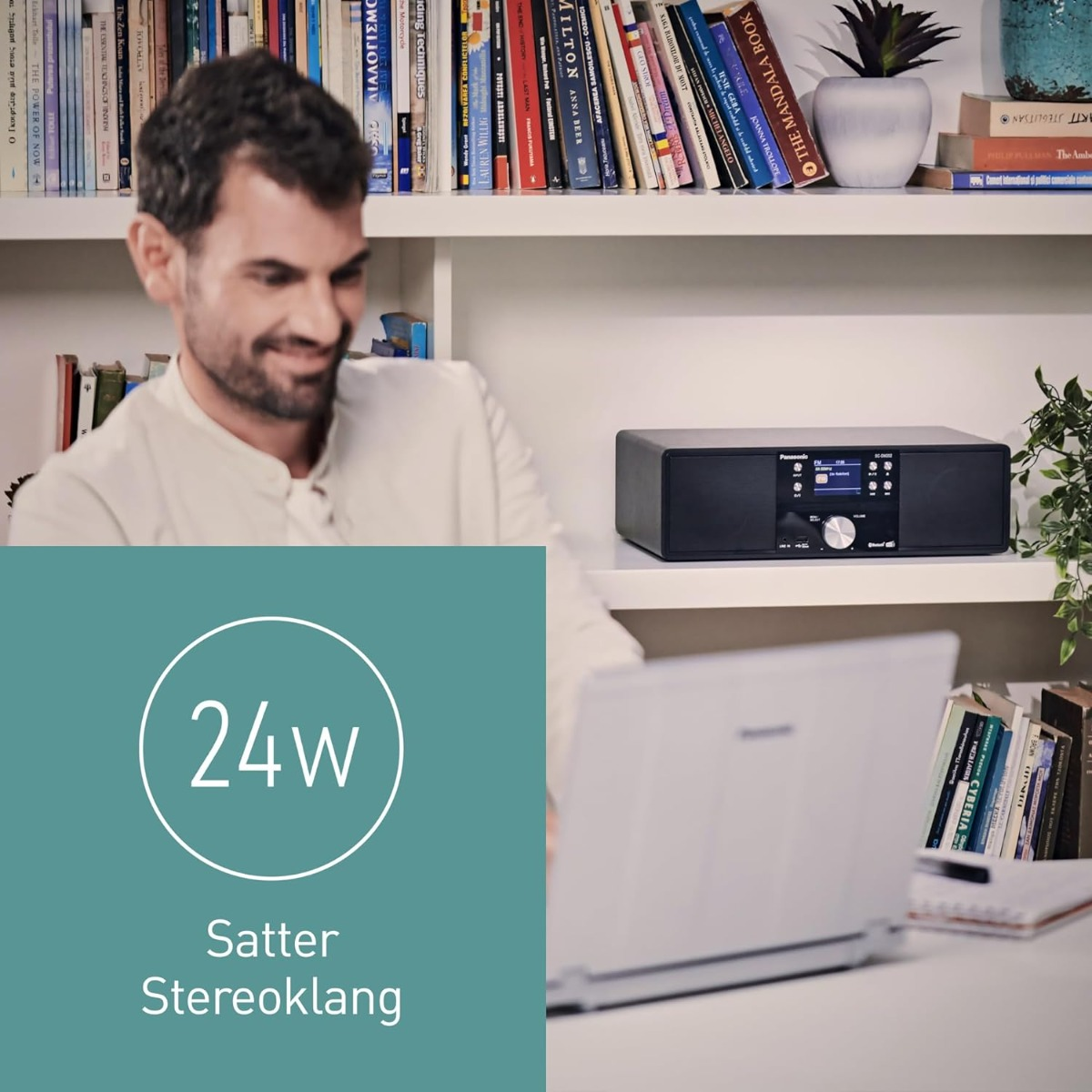 Mann lächelt in der Nähe einer Stereoanlage, Bücher in Regalen und ein Laptop. Ein Schild zeigt 24W Satter Stereoklang.