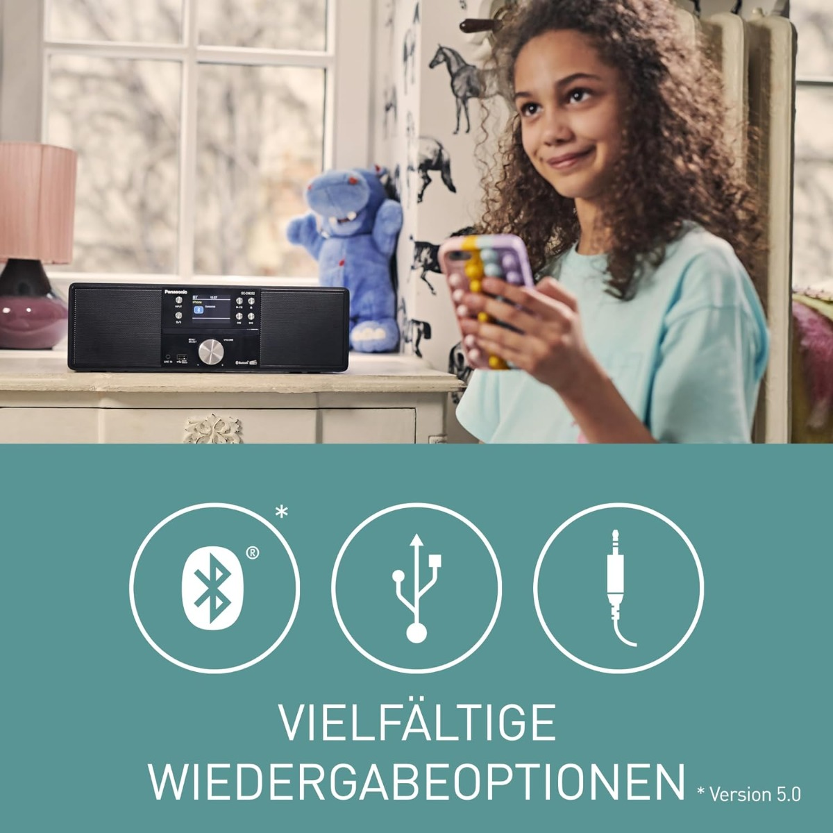 Ein Mädchen hält ein Telefon in der Nähe eines Lautsprechers. Ein blaues Nilpferd-Spielzeug und eine Lampe sind sichtbar. Text unten: 'VIELFÄLTIGE WIEDERGABEOPTIONEN'.