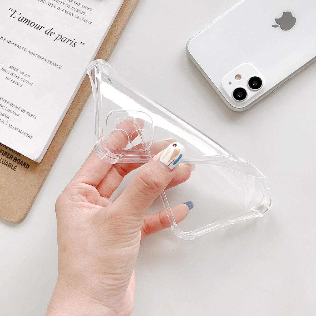 Una mano tiene una custodia trasparente per telefono. Sullo sfondo un iPhone bianco con doppia fotocamera. A sinistra un foglio con testo francese.