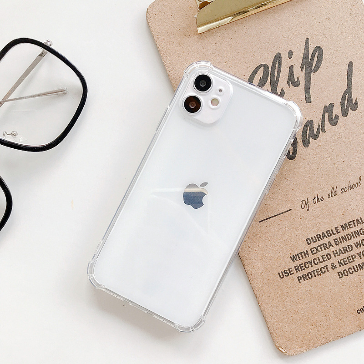 Uno smartphone bianco con una custodia trasparente è appoggiato su un fermacarte. Occhiali sono visibili. Lo sfondo è bianco.