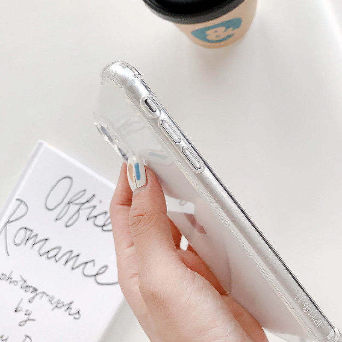 Una mano tiene una custodia trasparente per telefono. È visibile un libro intitolato 'Office Romance'. Sono presenti una tazza di caffè e una mano con nail art.