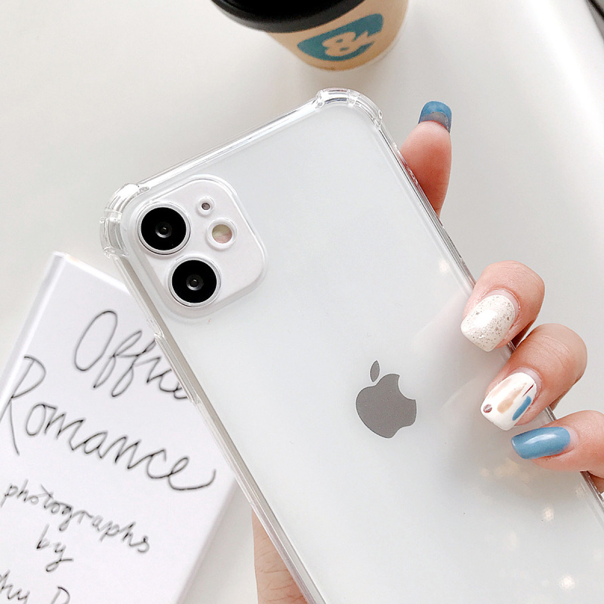 Una mano tiene un iPhone bianco con una custodia trasparente. Una tazza di caffè e un libro sono sullo sfondo. Le unghie sono dipinte di blu e con glitter.