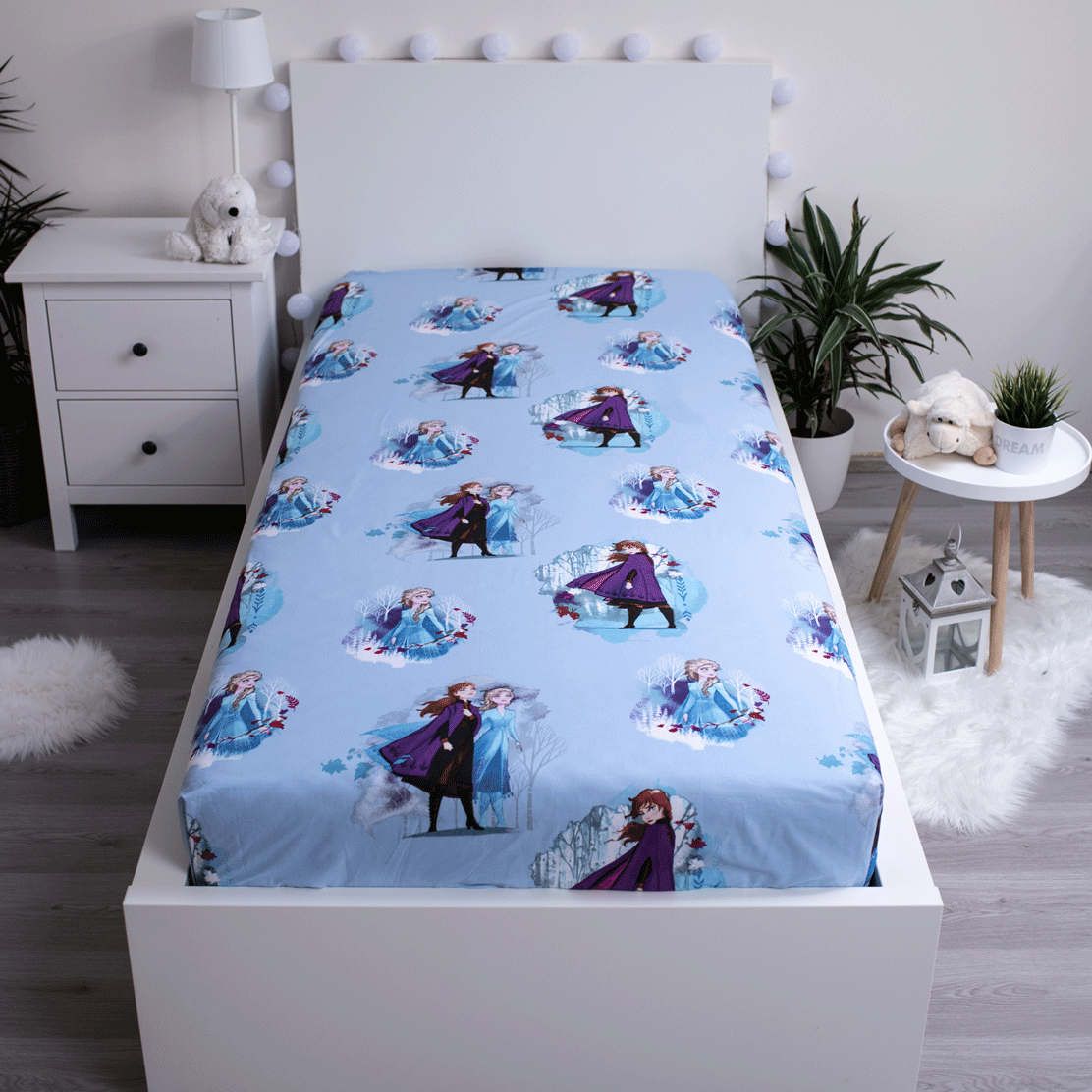 Ein Bett mit blauen Laken mit Frozen-Figuren. Ein weißes Kopfteil, Nachttisch und Pflanzen vervollständigen die Szene.