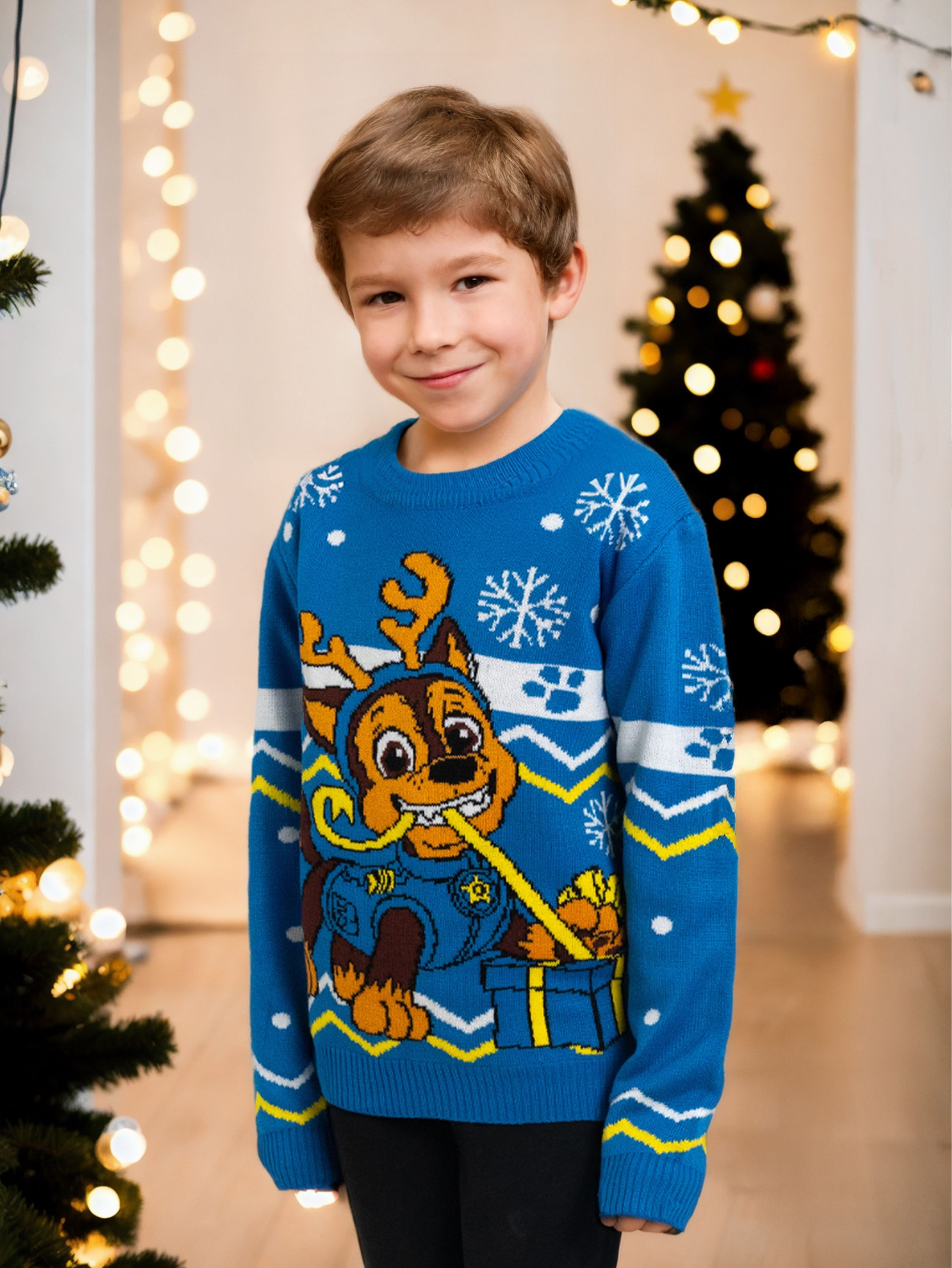 Ein Junge in einem blauen Pullover mit Paw Patrol-Design steht vor einem Weihnachtsbaum mit Lichtern.
