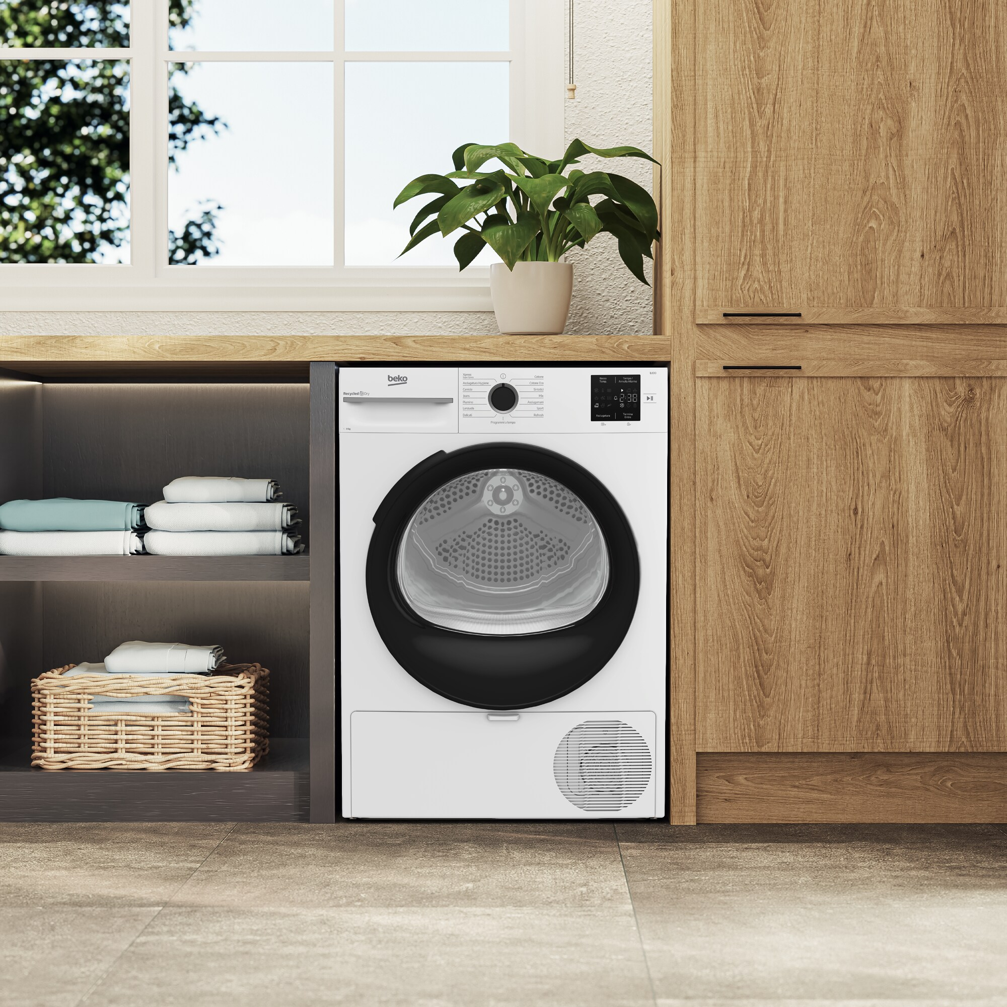Un'asciugatrice Beko bianca è accanto a un mobile in legno. Una pianta è sopra e una finestra con alberi è visibile.