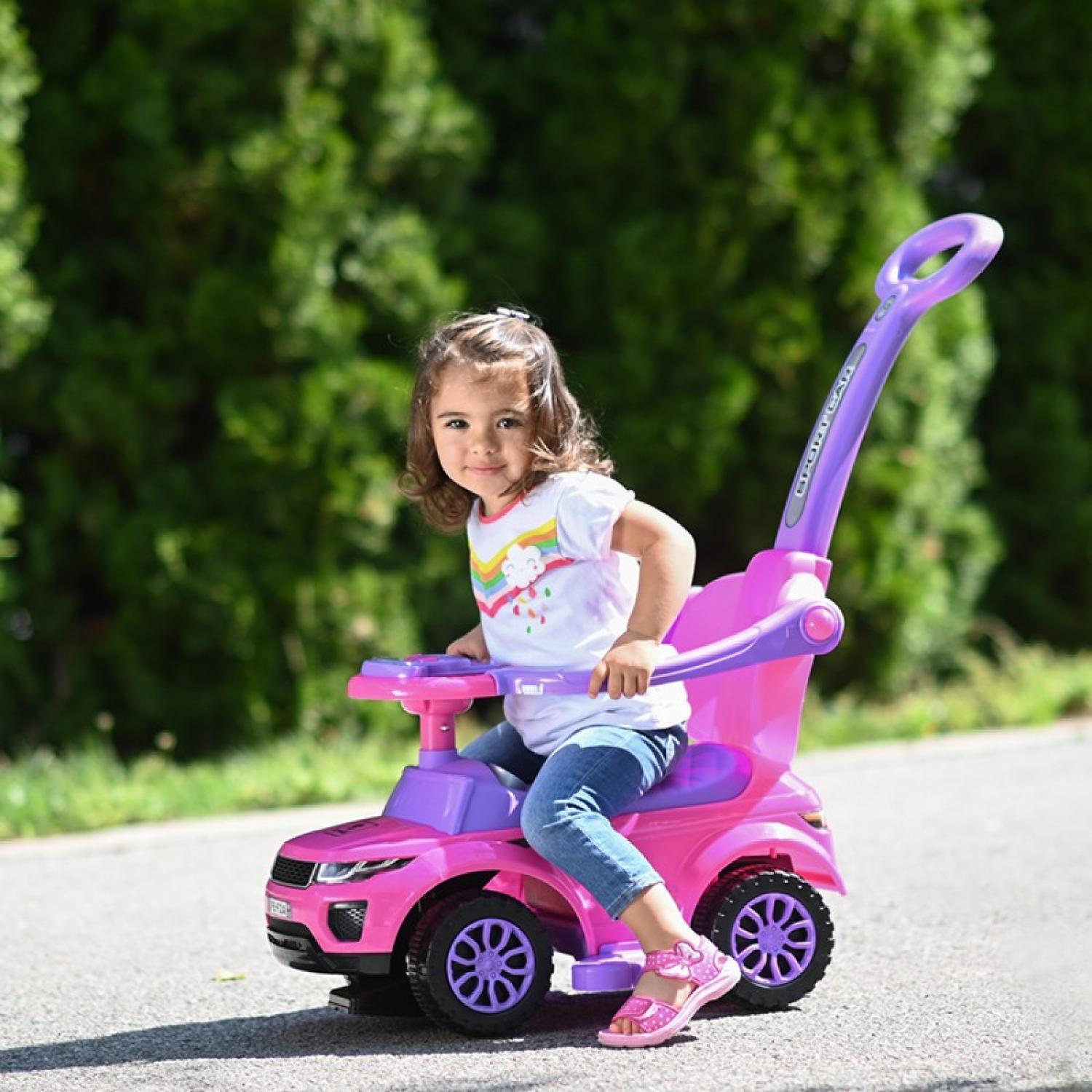 Una niña pequeña está sentada en un coche de juguete rosa. Lleva una camiseta blanca, vaqueros y sandalias rosas. El coche tiene ruedas moradas y un asa.