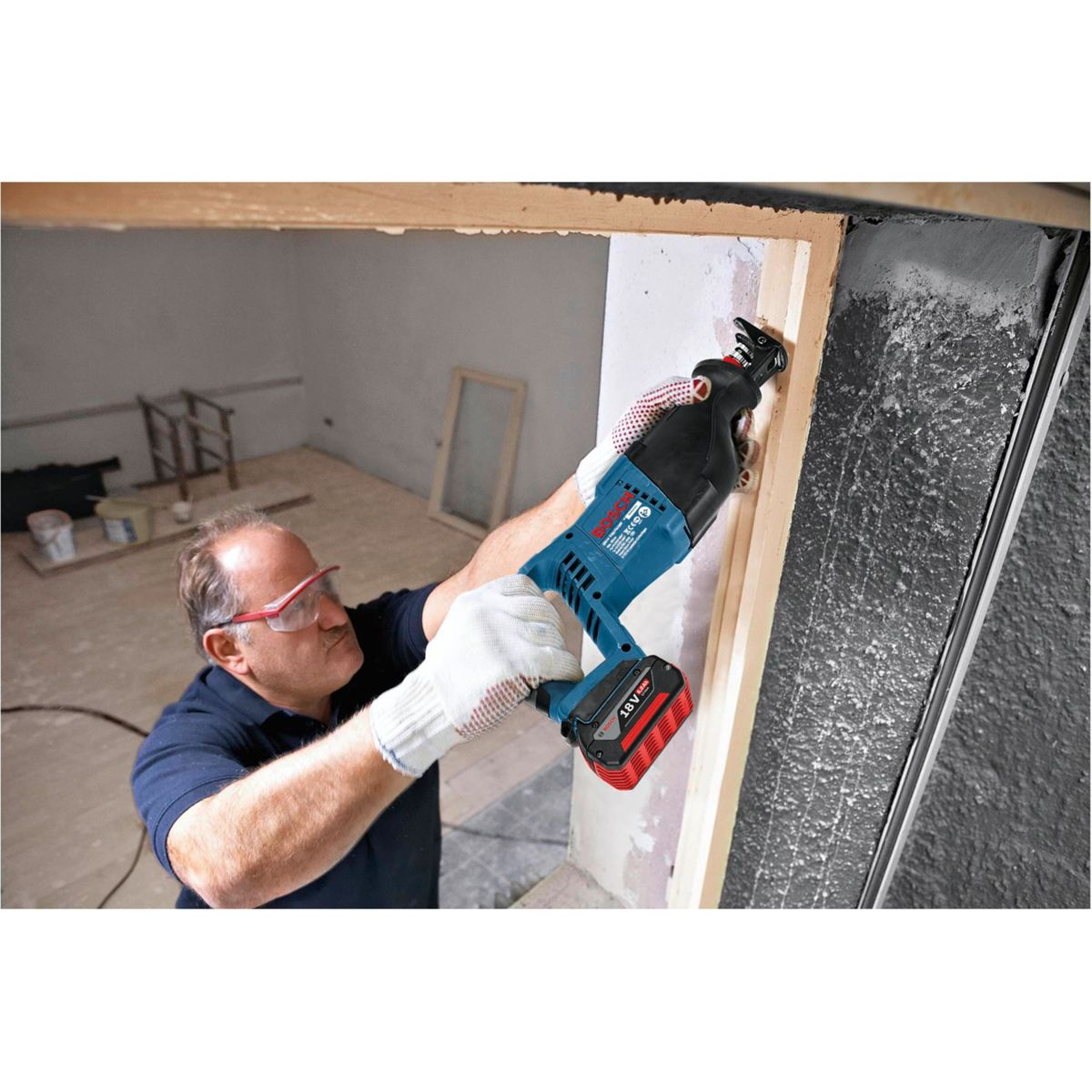 Mężczyzna używa niebiesko-czarnej piły szablastej do cięcia ramy drewnianej. Ma na sobie rękawice i okulary ochronne. Tłem jest pokój.