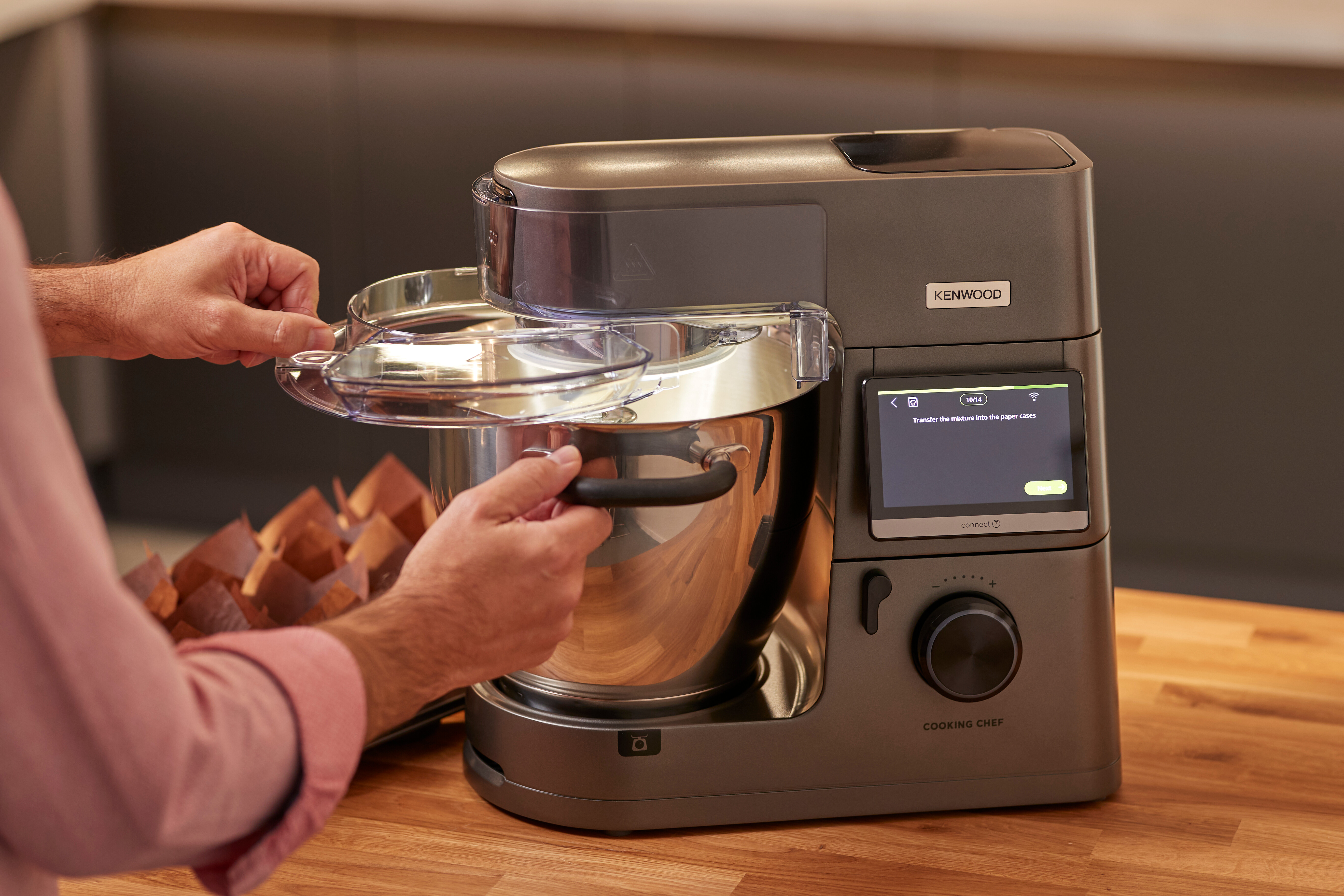 Eine Person, die einen Kenwood Cooking Chef benutzt. Das Gerät steht auf einer Holzoberfläche, mit Anweisungen auf dem Bildschirm.