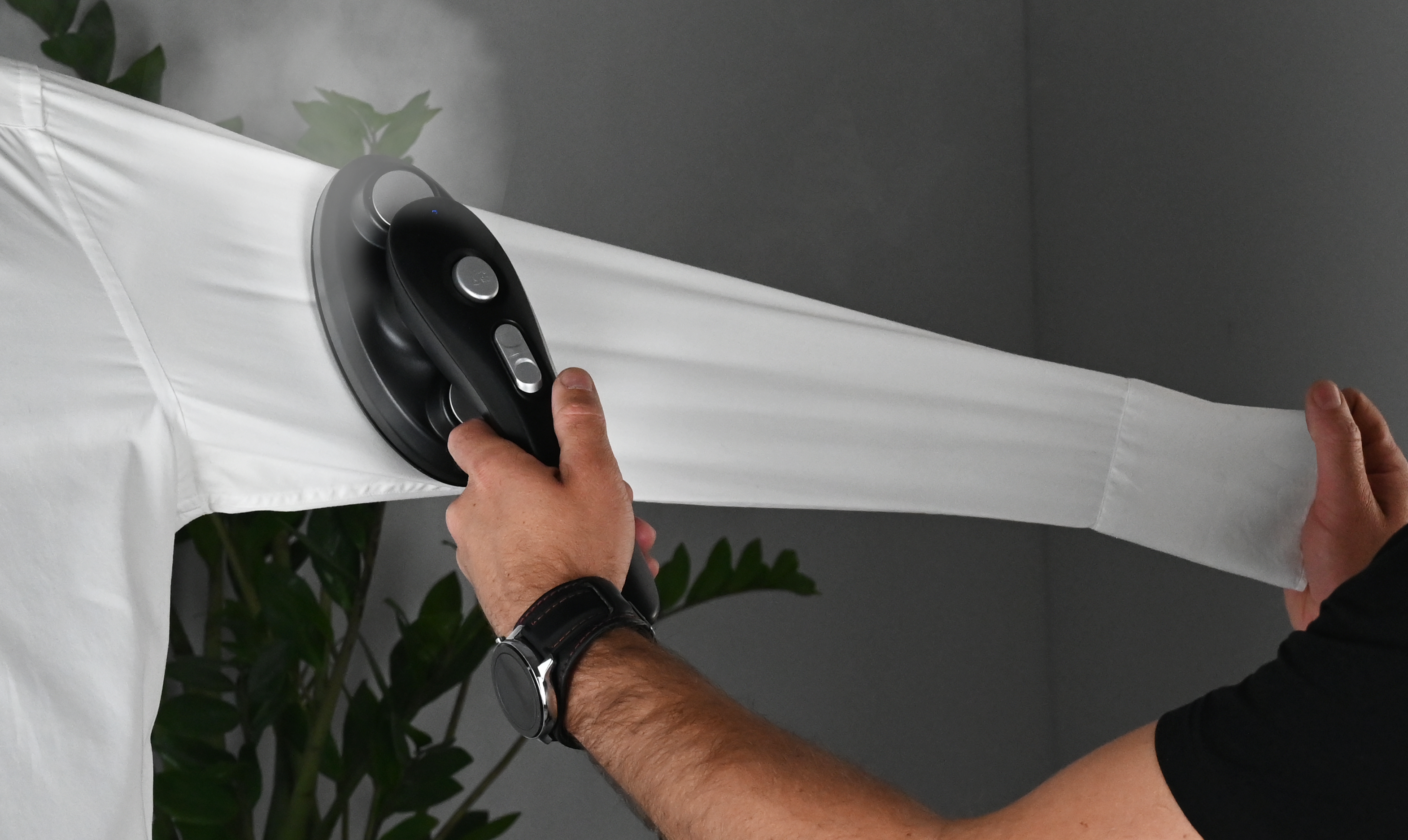 Osoba prasuje białą koszulę czarnym żelazkiem. Para jest widoczna. Tło to gładka ściana i kilka zielonych roślin.