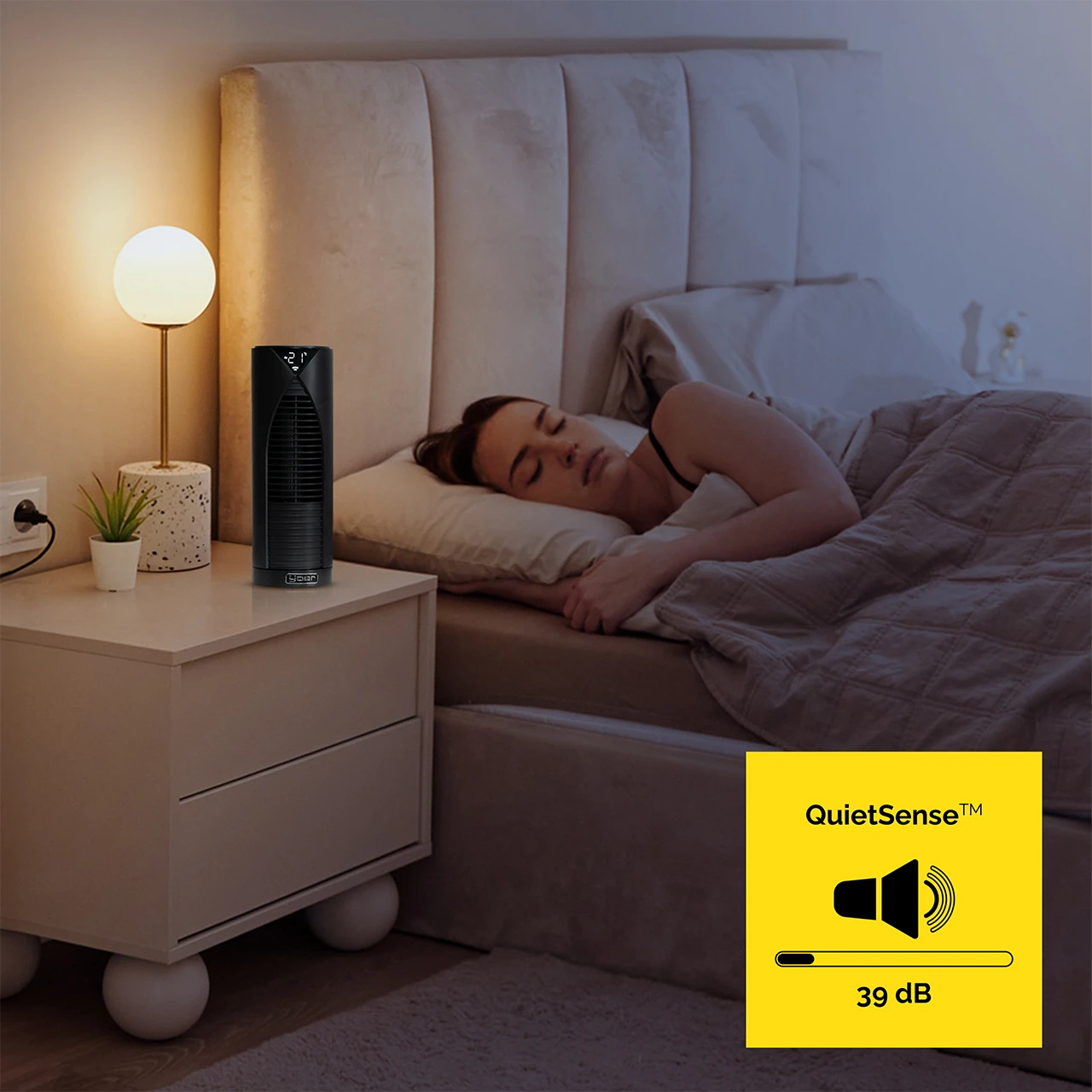 Kobieta śpi w sypialni. Czarny wentylator stoi na stoliku nocnym obok niej. Lampa i roślina również znajdują się na stoliku nocnym.