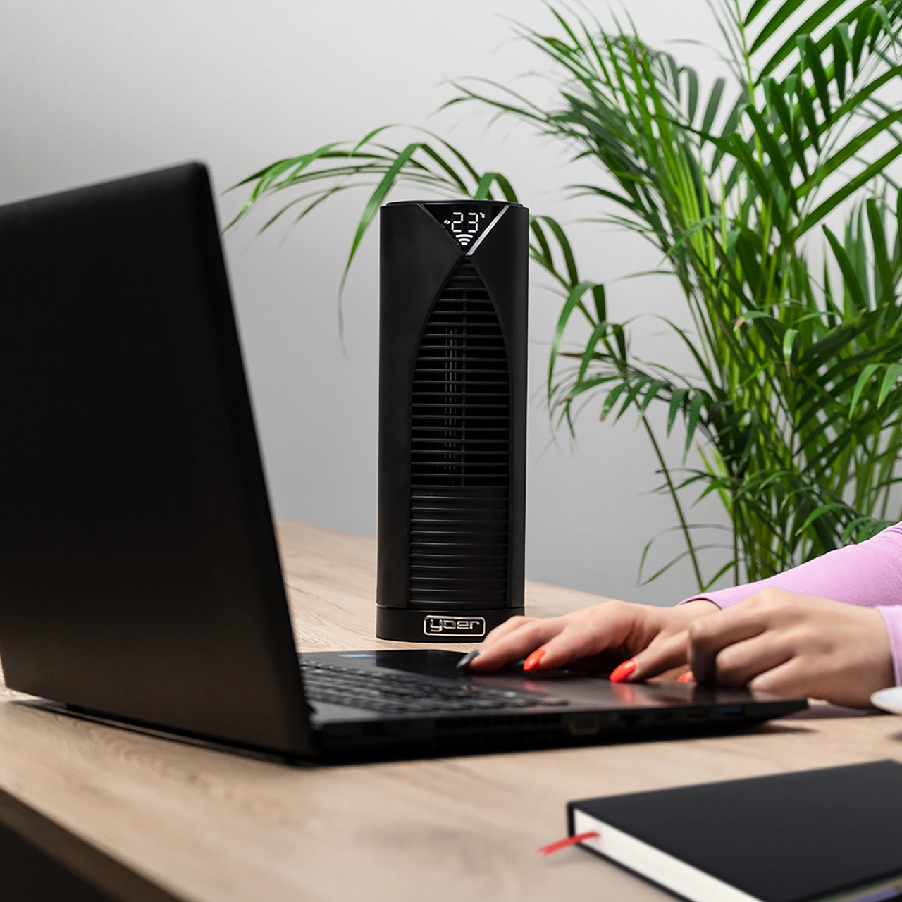 Kobieta używa laptopa na drewnianym biurku. Czarny wentylator stoi obok laptopa. Roślina jest w tle.