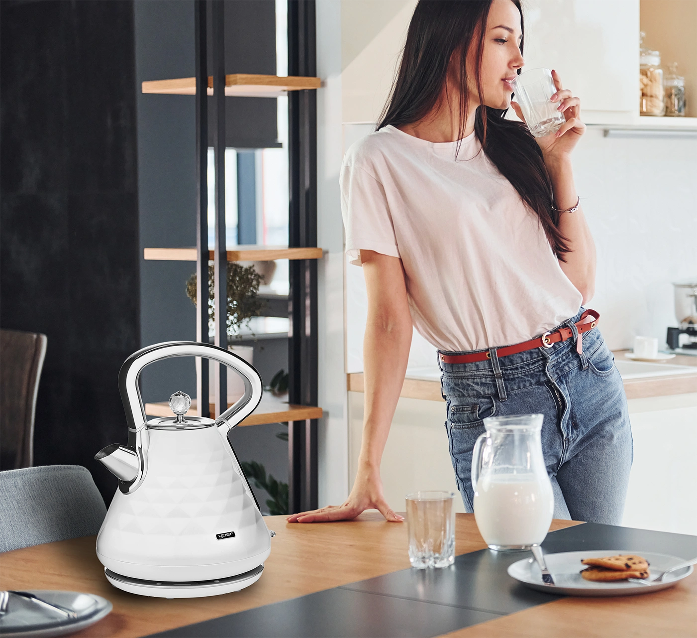 Kobieta pije wodę w kuchni. Biały czajnik na stole. Mleko i ciastka również na stole.