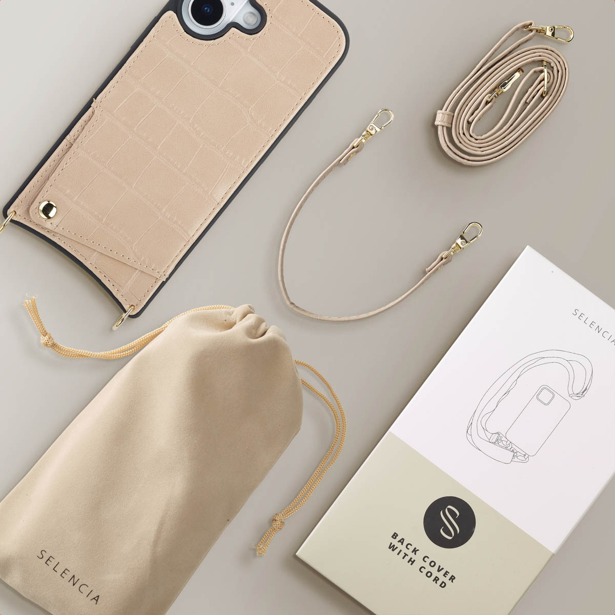 Une coque de téléphone beige avec une sangle, un sac et un dépliant sur une surface gris clair. La coque a un motif crocodile.