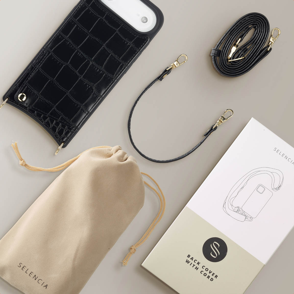 Een zwarte telefoonhoes met een riem, een bijpassende riem, een beige zakje en een doos met een telefoonhoes-illustratie liggen op een grijs oppervlak.