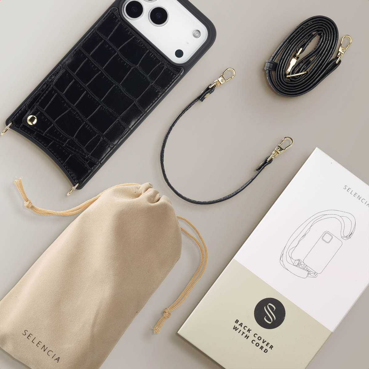 Une vue aérienne montre un étui de téléphone noir, une sangle et une pochette beige, avec l'emballage du produit. Le tout sur une surface gris clair.