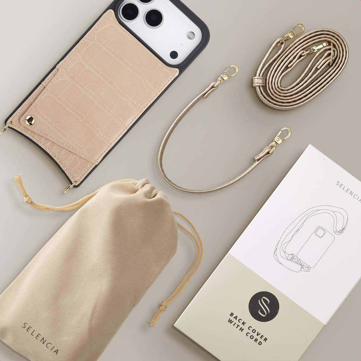 Een beige telefoonhoesje met een band, een zakje met trekkoord, een gouden band en een doos zijn gerangschikt op een lichtgrijze ondergrond.