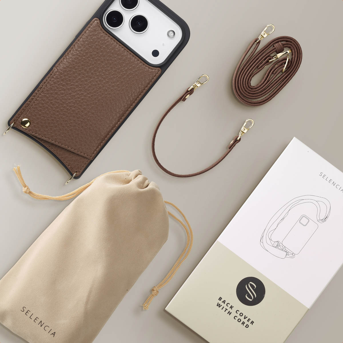 Bruine leren telefoonhoes met riem, beige zakje en witte doos op een grijze ondergrond. De hoes heeft gouden accenten.