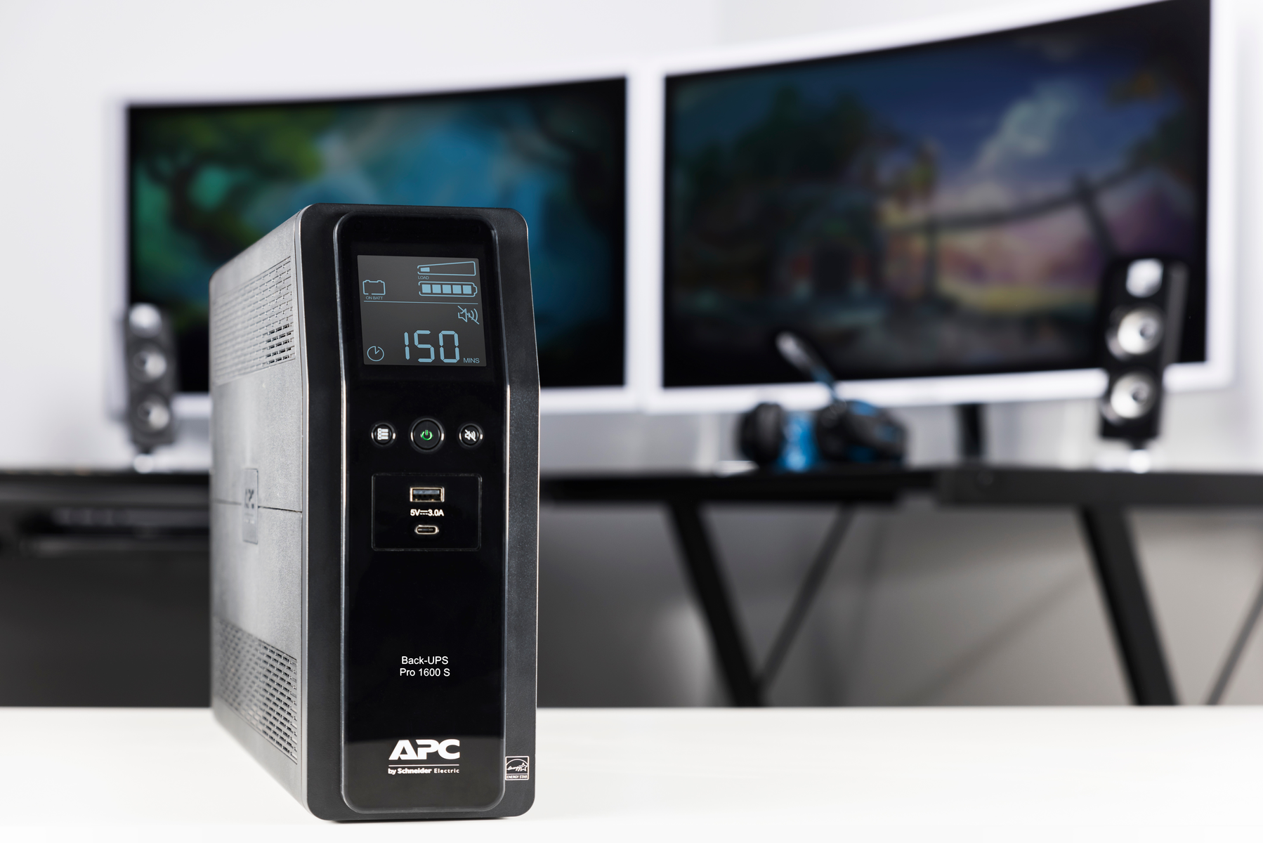 Un APC Back-UPS Pro 1600 S nero è su una scrivania bianca. Due monitor e altoparlanti sono sfocati sullo sfondo.