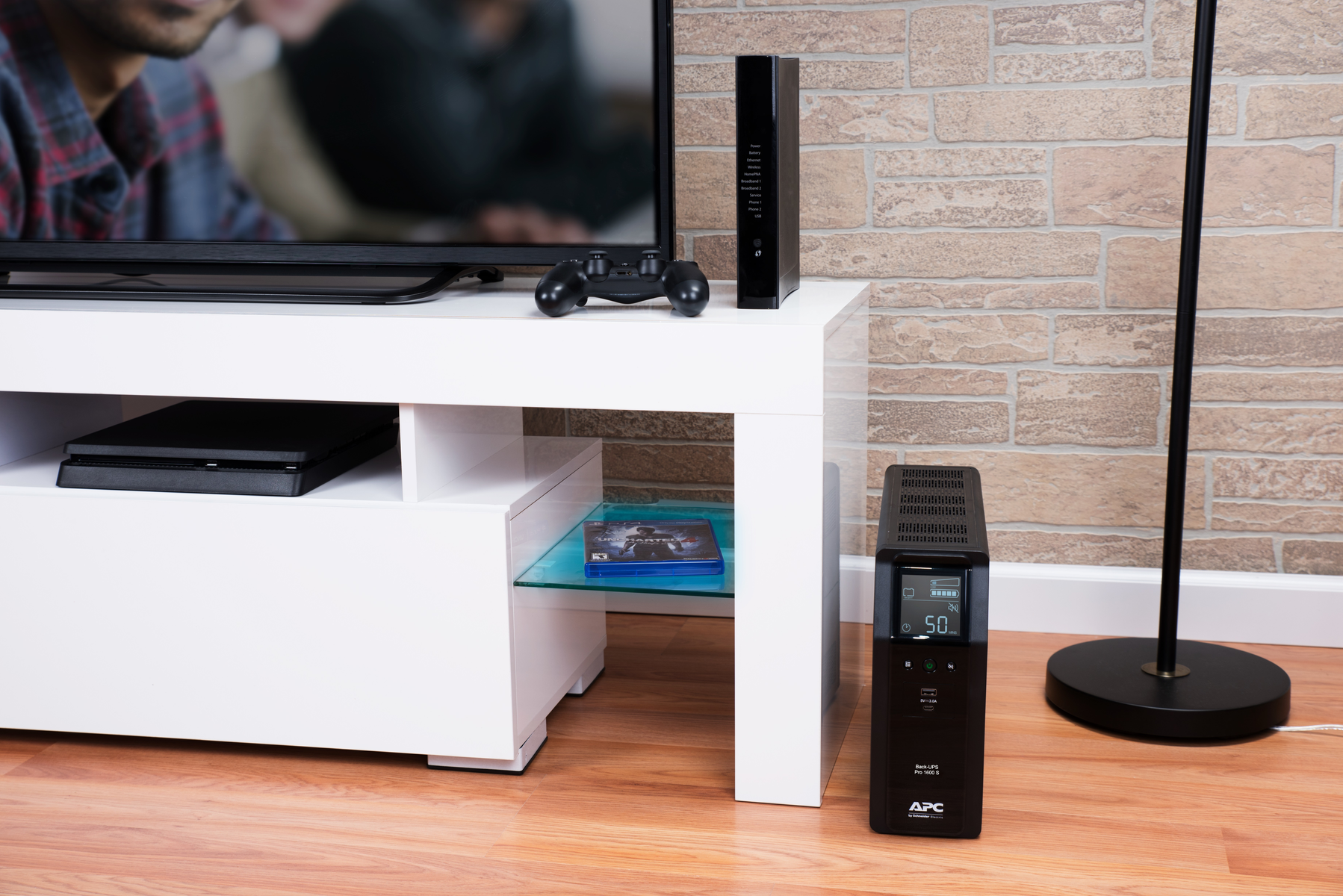 Soggiorno con TV, tavolo bianco, console di gioco, modem e UPS. Visibili un muro di mattoni e pavimento in legno.