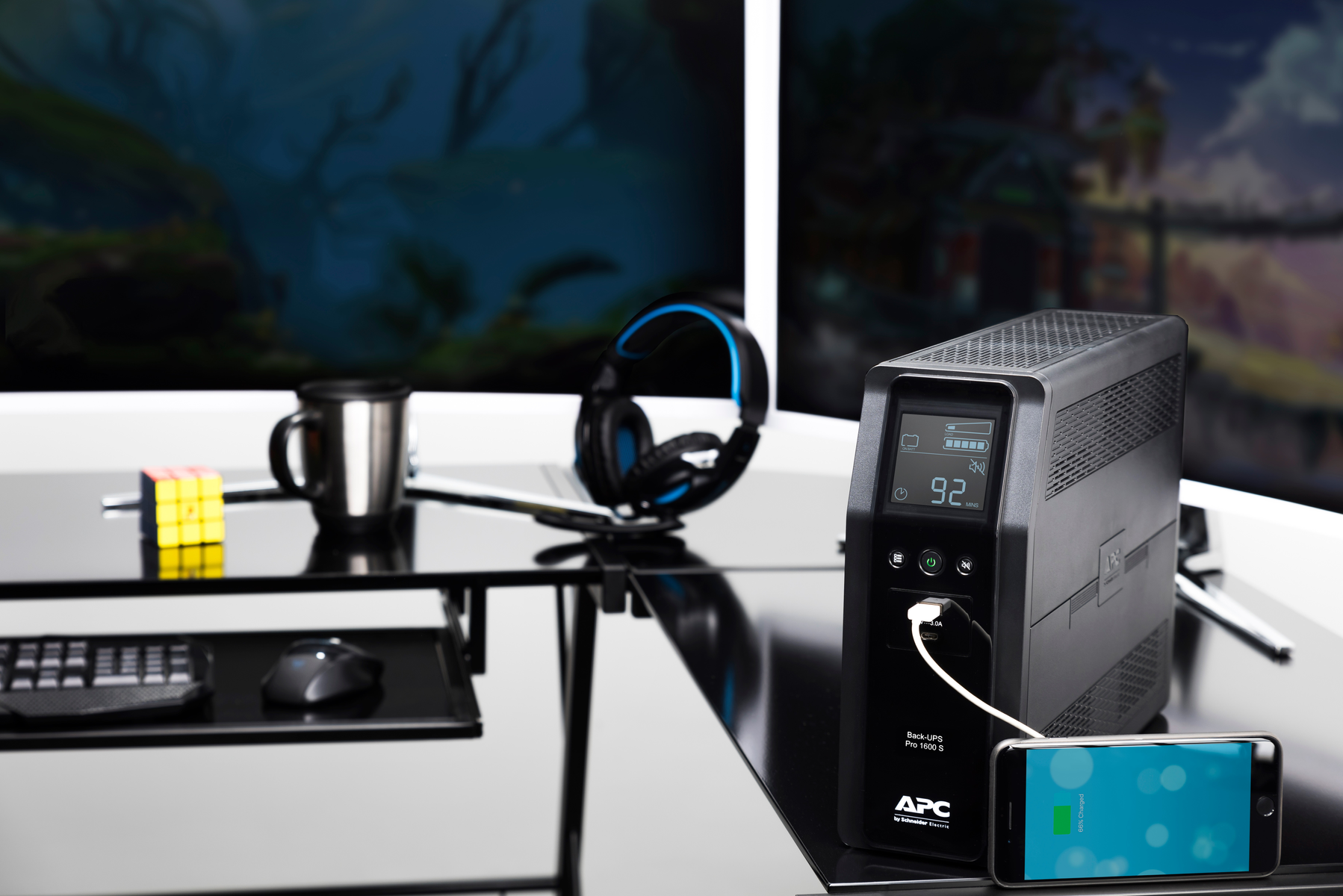 Un UPS nero con un telefono in carica, cuffie, una tazza e un cubo di Rubik su una scrivania. Lo sfondo include monitor.