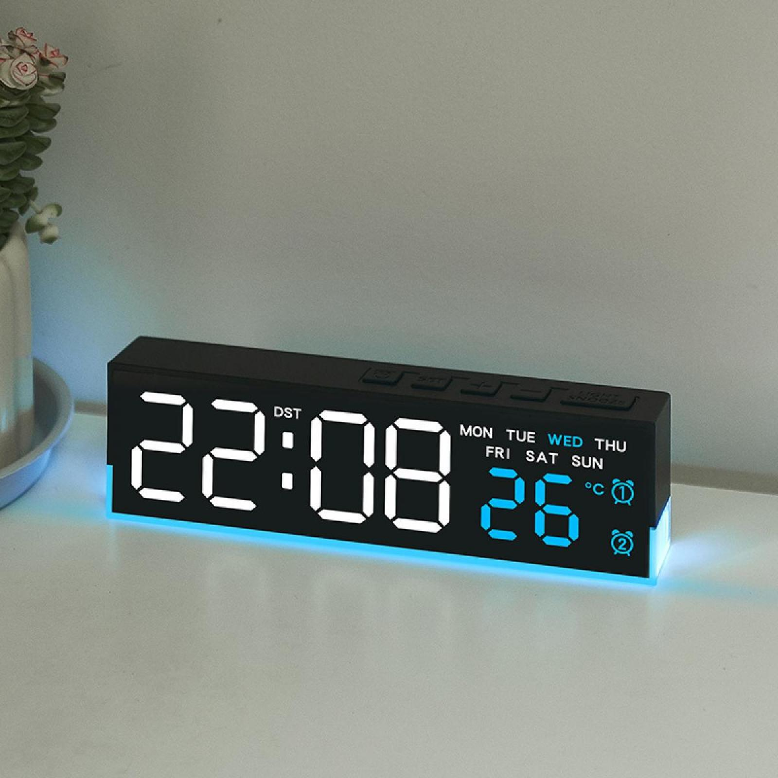 Eine Digitaluhr zeigt 22:08 und 26 Grad an. Sie steht auf einer weißen Oberfläche mit einer Pflanze im Hintergrund.