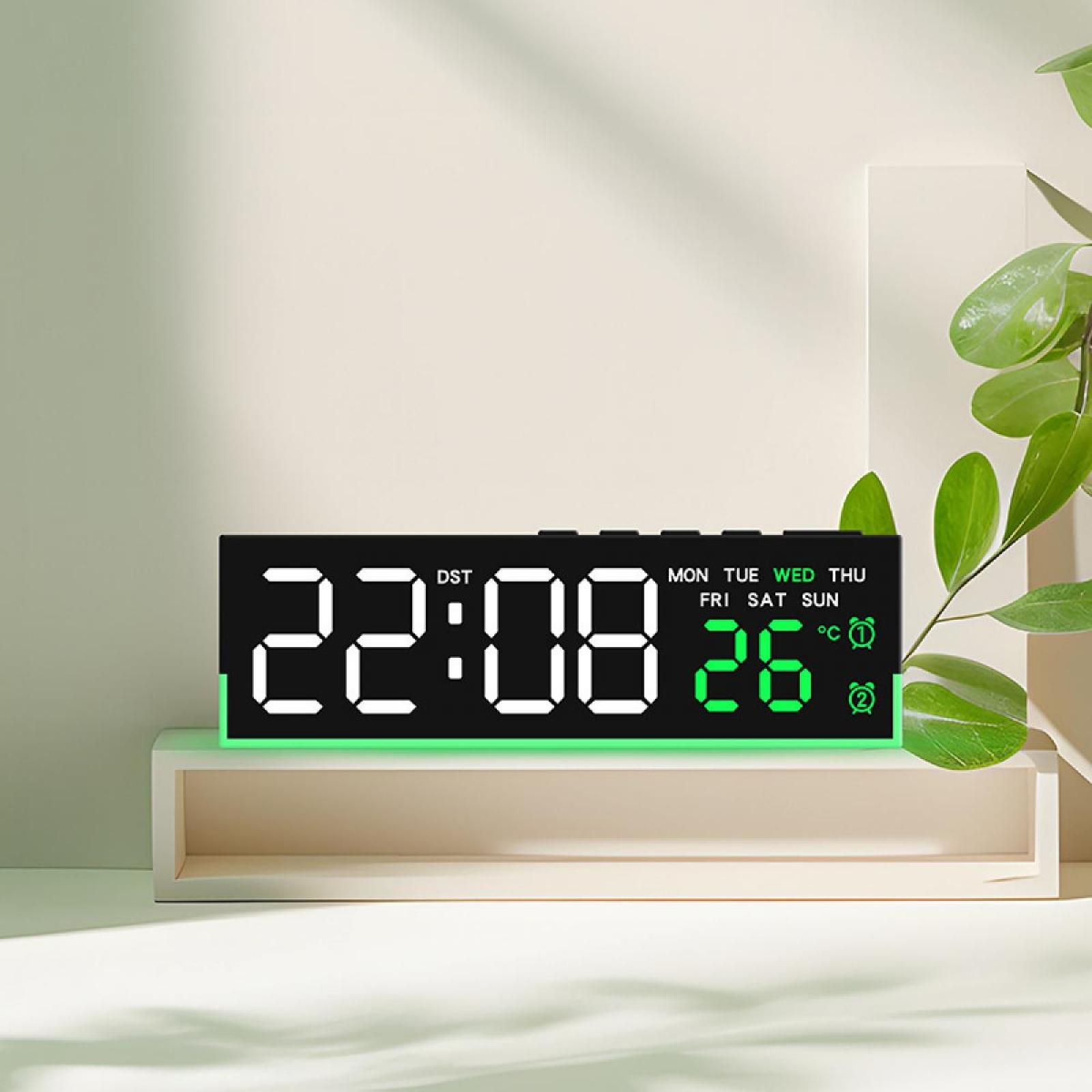 Digitaluhr mit der Anzeige 22:08, 26 Grad, Tag und Uhrzeit. Liegt auf einem weißen Regal, mit grüner Pflanze im Hintergrund.