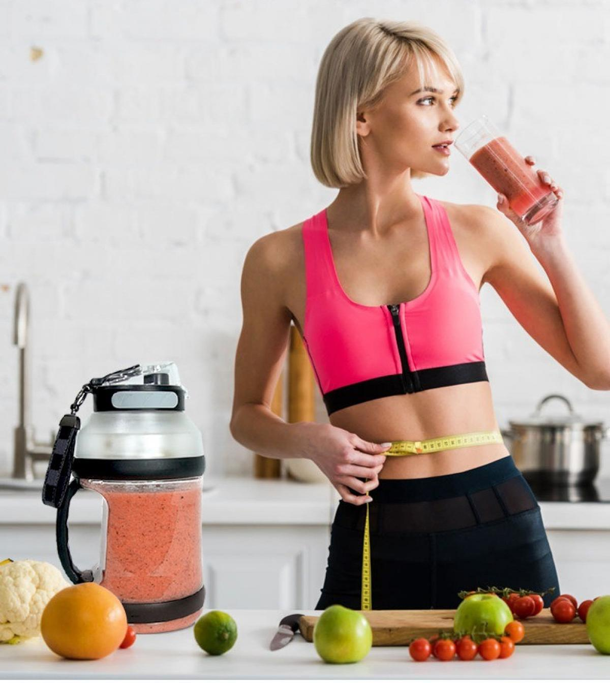 Frau in rosa Oberteil trinkt Smoothie, misst Taille. Smoothie-Glas, Früchte, Gemüse auf dem Tisch.