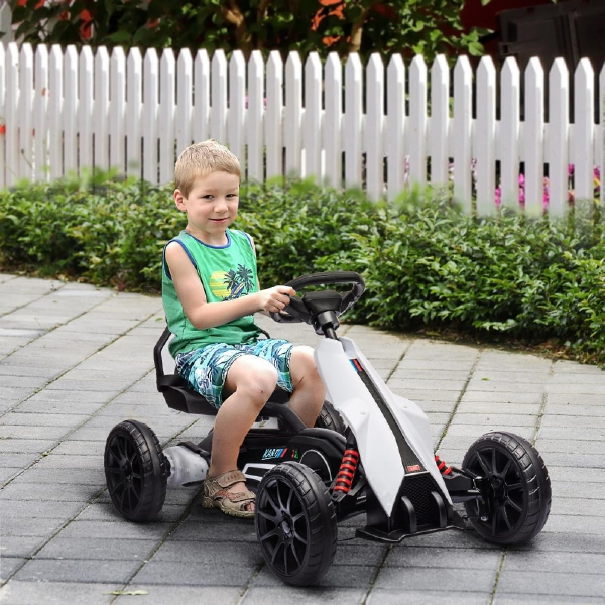 Ein Junge sitzt in einem weißen und schwarzen Go-Kart. Er lächelt und trägt ein grünes Hemd und blaue Shorts. Ein weißer Zaun und grüne Büsche sind hinter ihm.