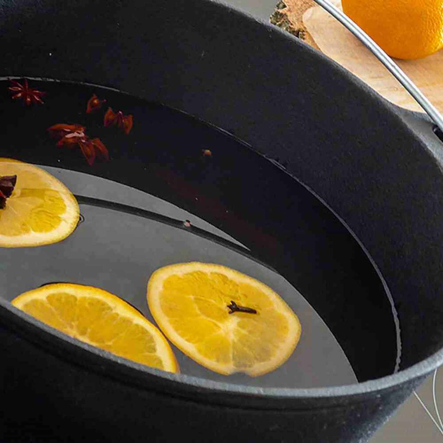 Ein schwarzer Topf mit Wasser und Orangenscheiben, Sternanis und Nelken. Im Hintergrund eine Orange und ein Holzbrett.