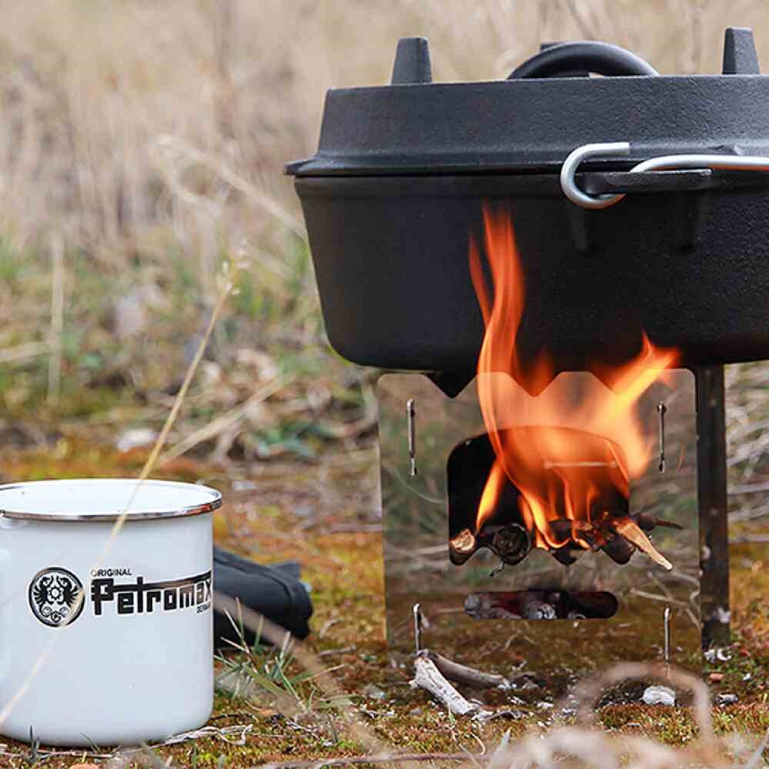 Ein Lagerfeuer mit einem schwarzen Topf darauf. Auf der linken Seite befindet sich ein weißer Becher mit Logo. Der Hintergrund ist ein verschwommenes Feld.
