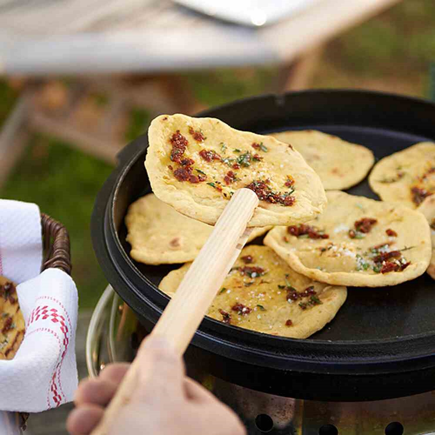 Ręka używa szczypiec do podniesienia podpłomyka z patelni. Inne podpłomyki są na patelni. Koszyk z tkaniną znajduje się w tle.