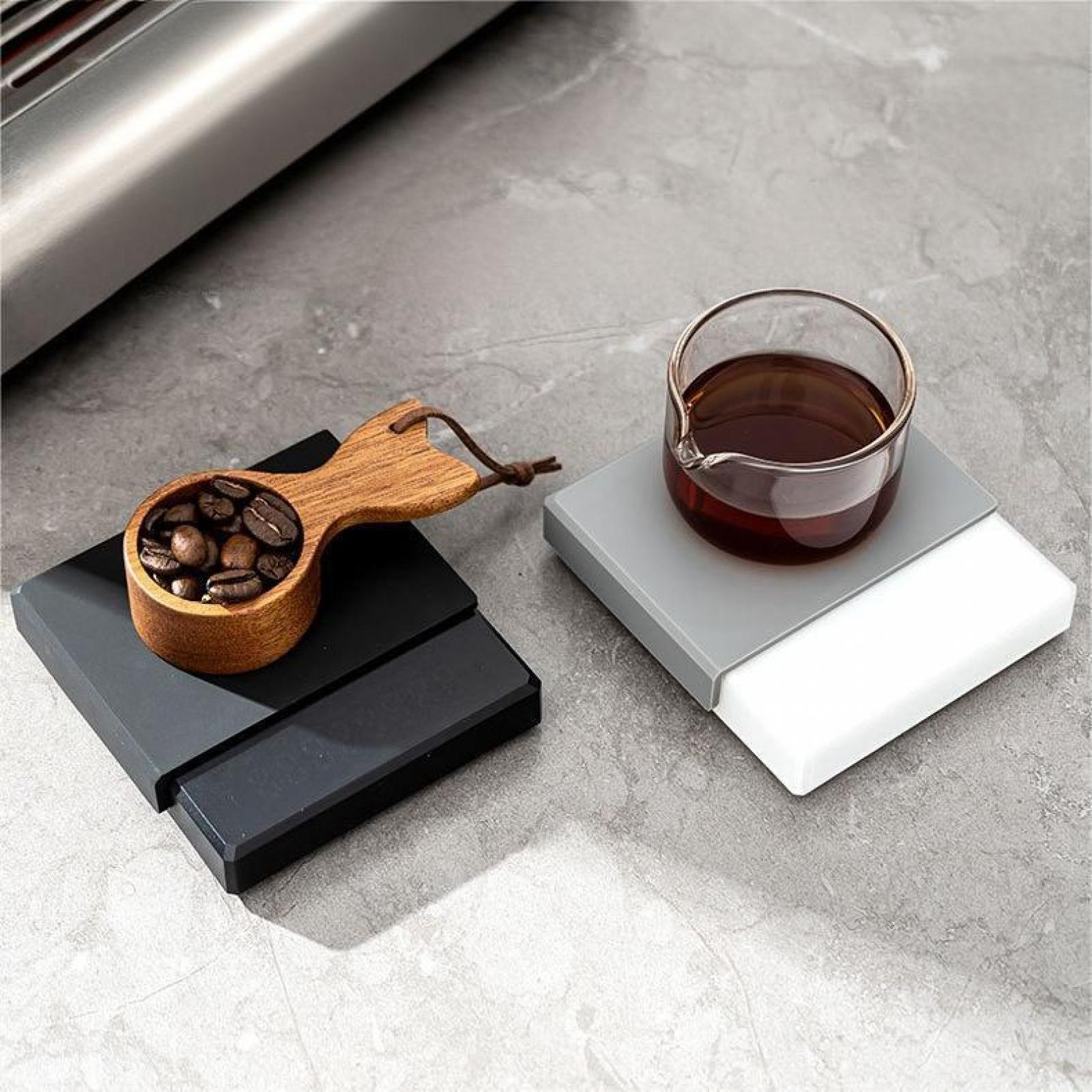 Ein hölzerner Löffel mit Kaffeebohnen neben einem Glas brauner Flüssigkeit auf einem grau-weißen Untersetzer.