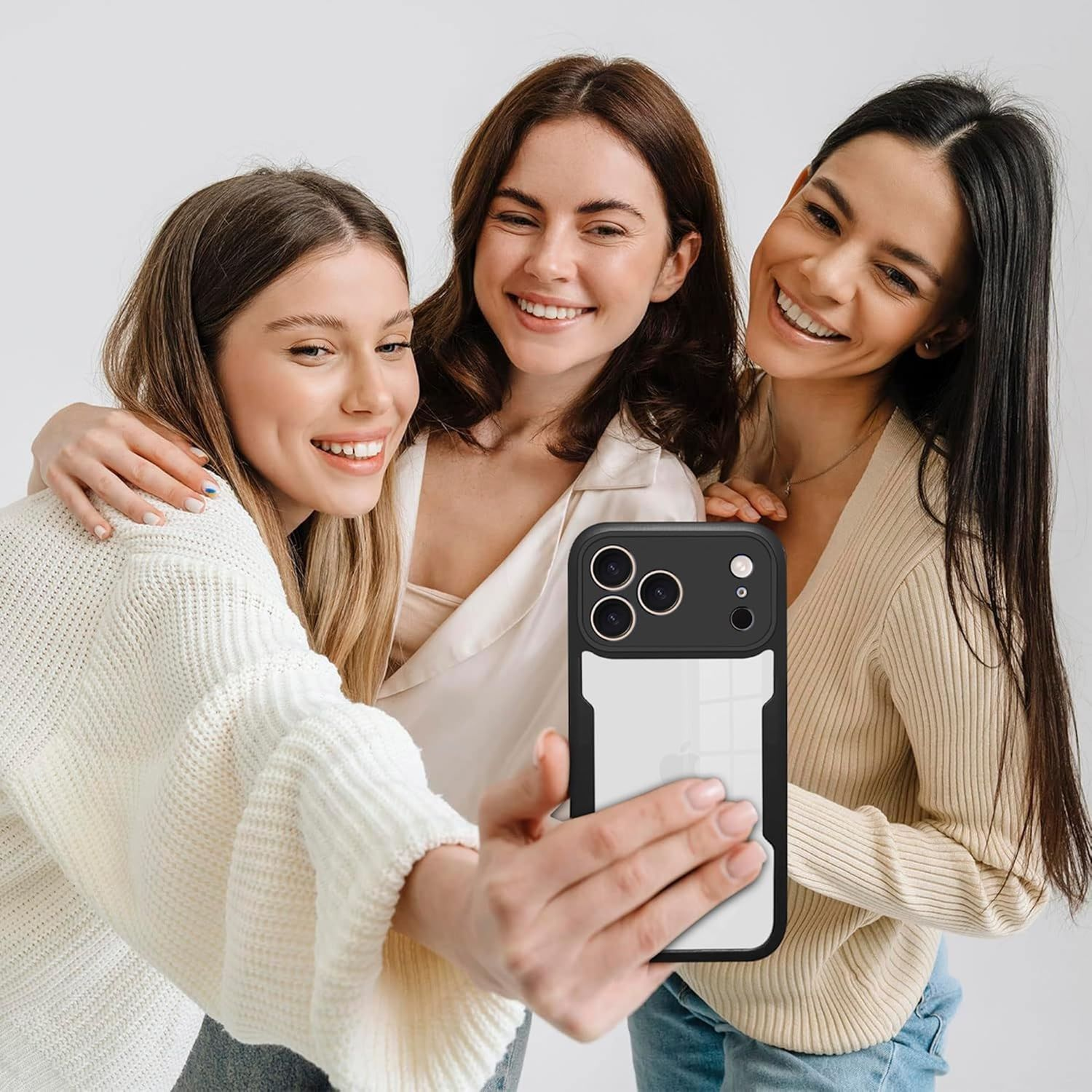 Drei lächelnde Frauen machen ein Selfie. Eine hält das Telefon, andere haben Arme umeinander. Weißer Hintergrund.