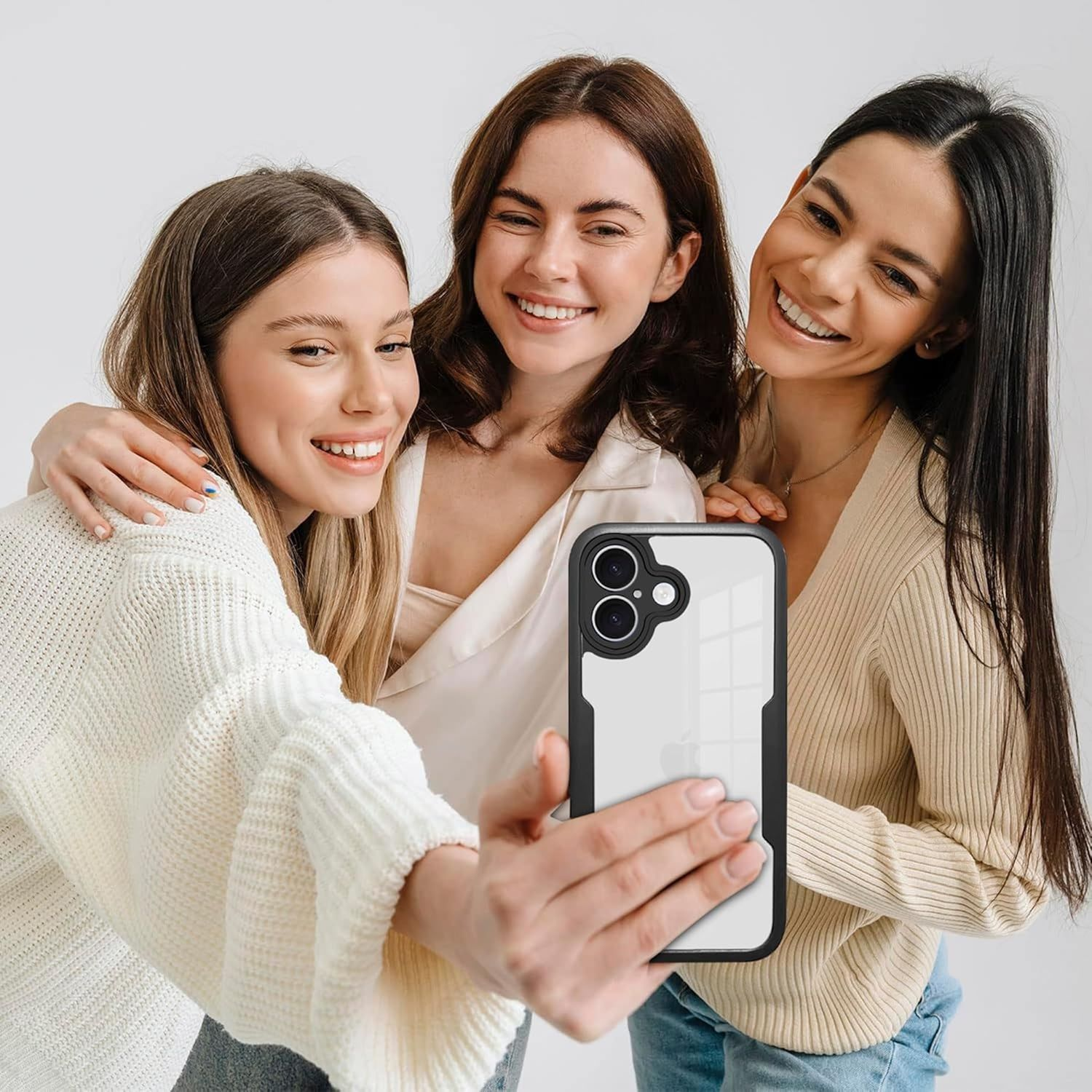 Drei lächelnde Frauen machen ein Selfie. Eine hält das Telefon. Alle sind eng beieinander, weißer Hintergrund.