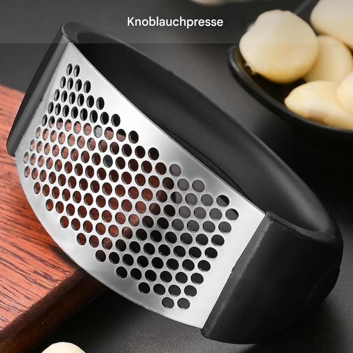 Eine Knoblauchpresse mit silberner Metallpresse und schwarzem Griff. Knoblauchzehen befinden sich in einer schwarzen Schüssel im Hintergrund.