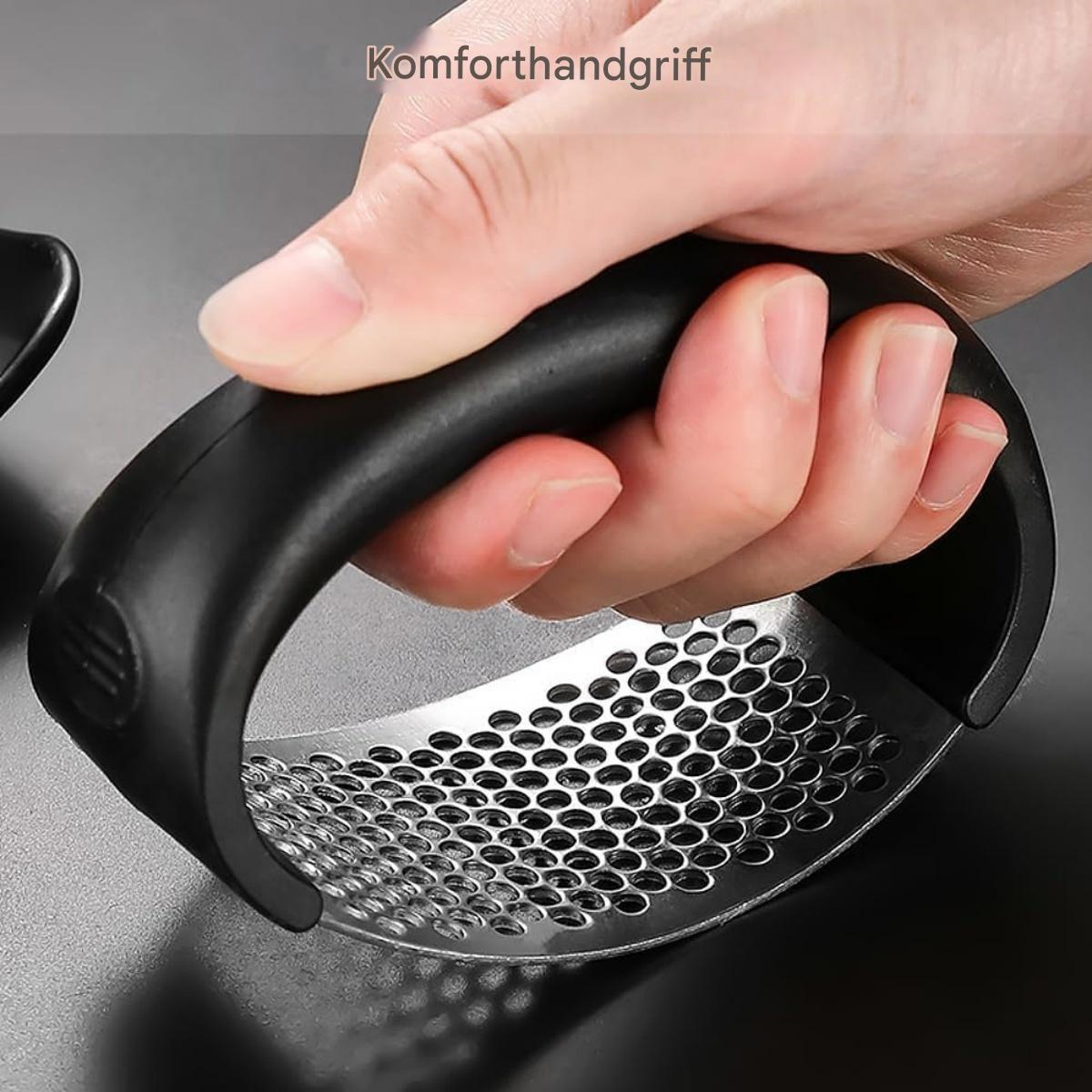 Eine Hand, die eine schwarze Knoblauchpresse greift. Die Presse hat eine silberne, perforierte Basis. Der Hintergrund ist eine dunkle Oberfläche.