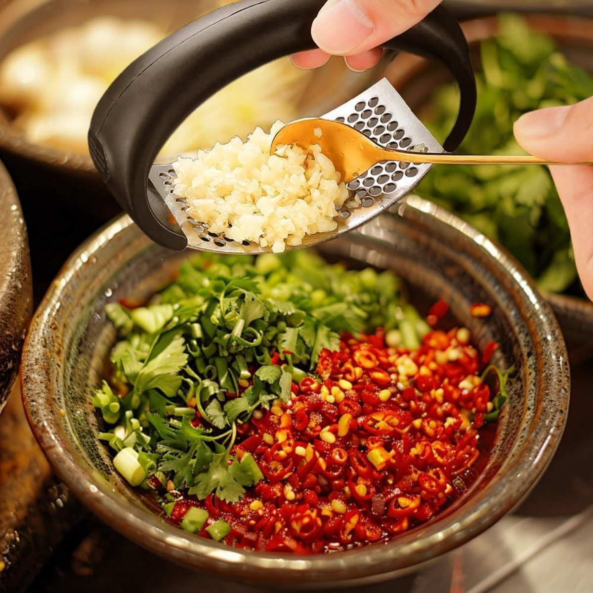 Eine Hand drückt Knoblauch durch eine Reibe in eine Schüssel mit gehackten Chilis und Kräutern. Ein goldener Löffel hält den Knoblauch.