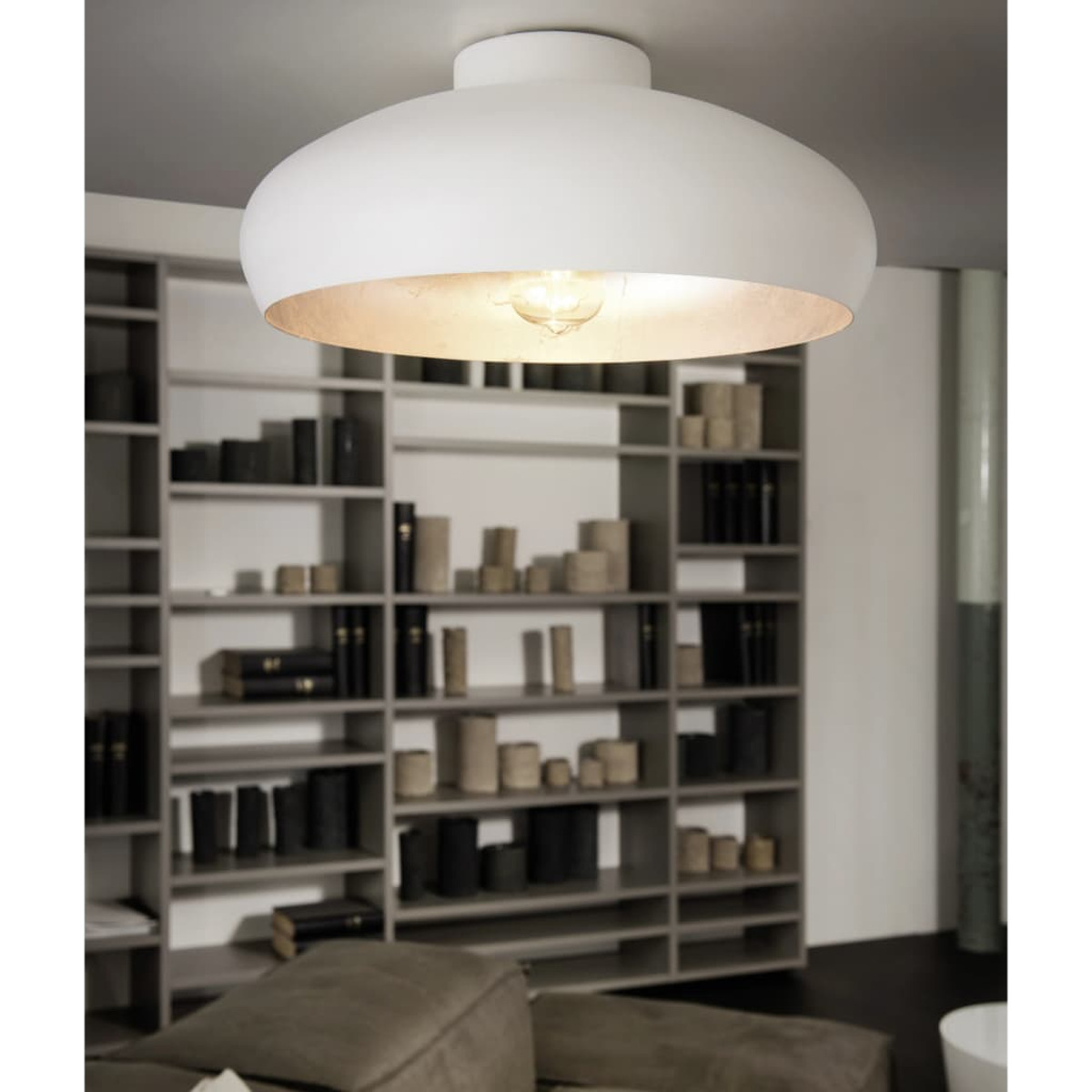 Eine weiße Deckenlampe mit einer brennenden Glühbirne hängt über einem Bücherregal mit Büchern und Dekorationsartikeln. Im Vordergrund befindet sich ein Sofa.