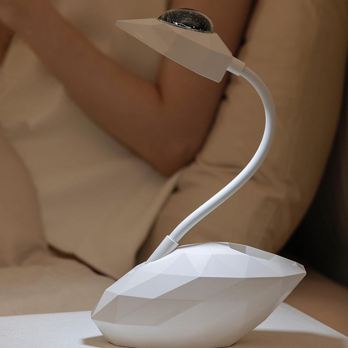 Eine weiße, geometrische Schreibtischlampe steht auf einem Tisch. Dahinter sitzt eine Person. Die Lampe hat einen flexiblen Hals und ein kleines Licht.