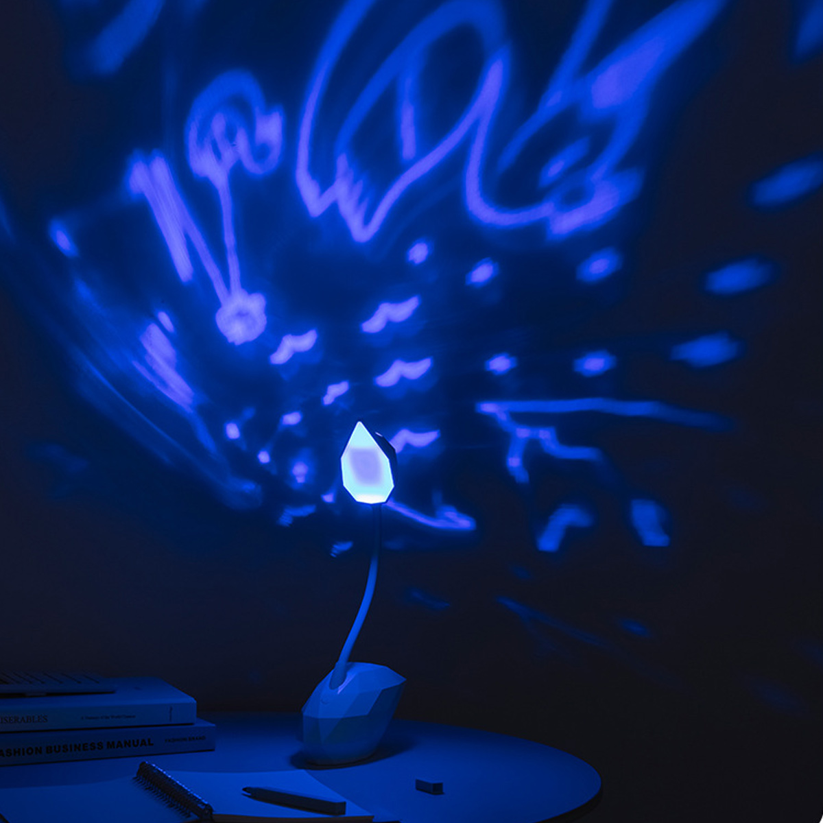 Eine blau beleuchtete Lampe in Form einer Blume steht auf einem Tisch. Abstrakte Formen werden an die Wand projiziert. Bücher und ein Stift sind ebenfalls sichtbar.