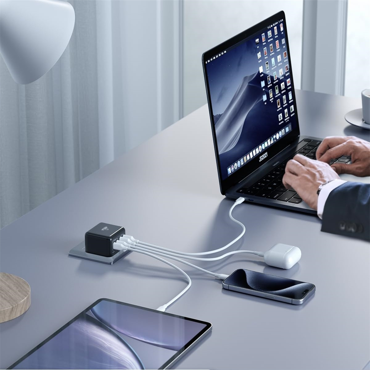 Laptop, Tablet und Telefon sind mit einem Netzteil verbunden. Eine Person tippt auf dem Laptop. Weiße Kabel verbinden Geräte.